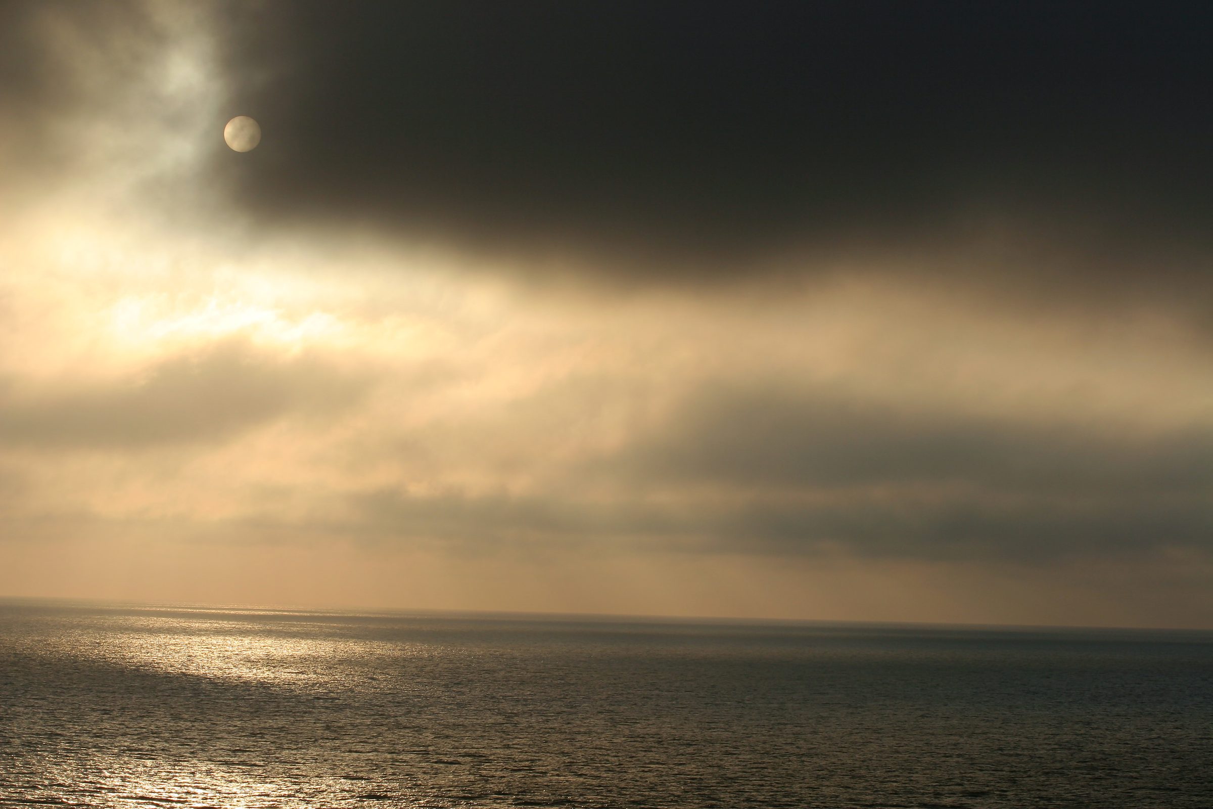 Golden sun behind dark clouds reflecting on a calm open sea.
