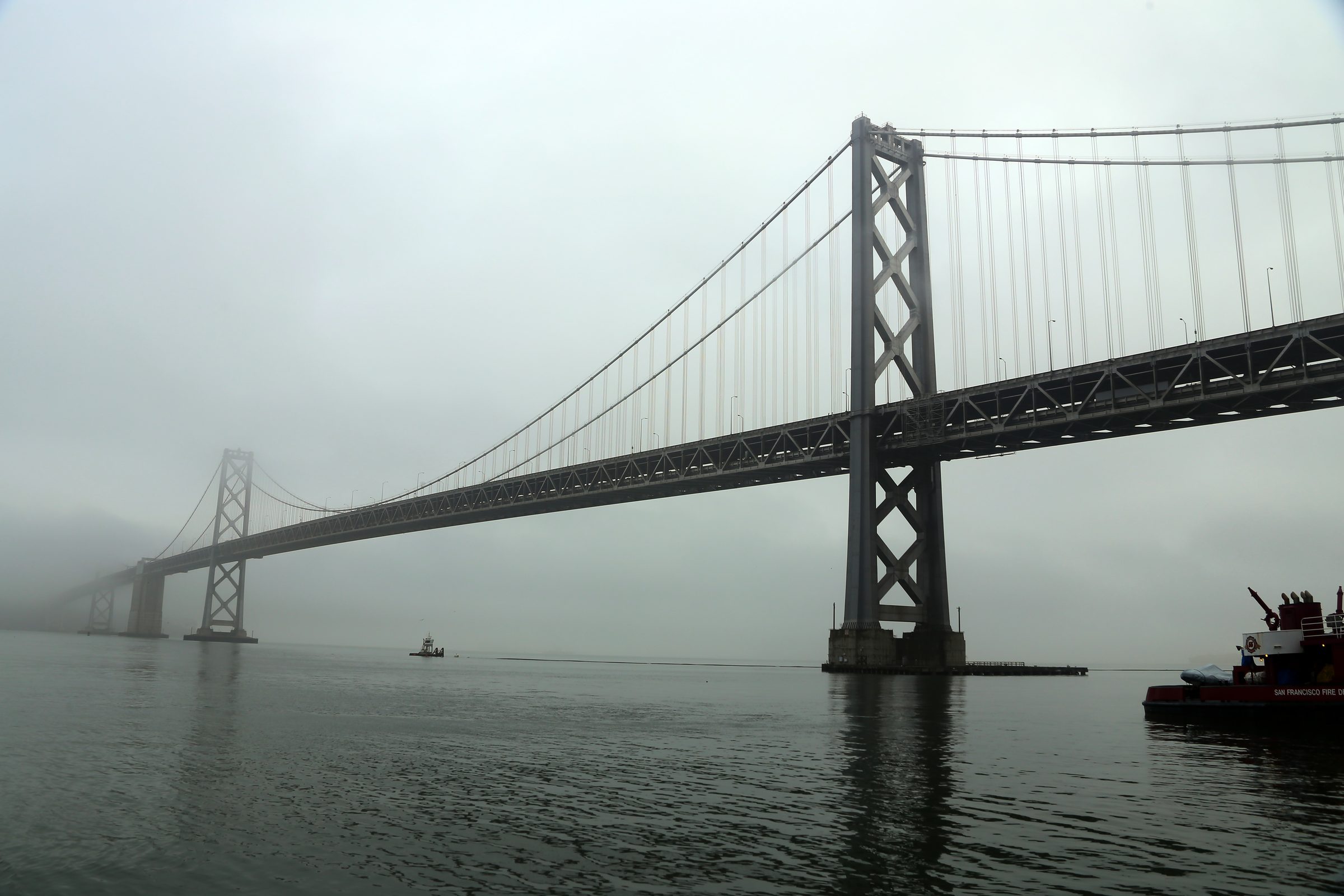 Bay Bridge X-bracing disappearing into fog over calm grey water.