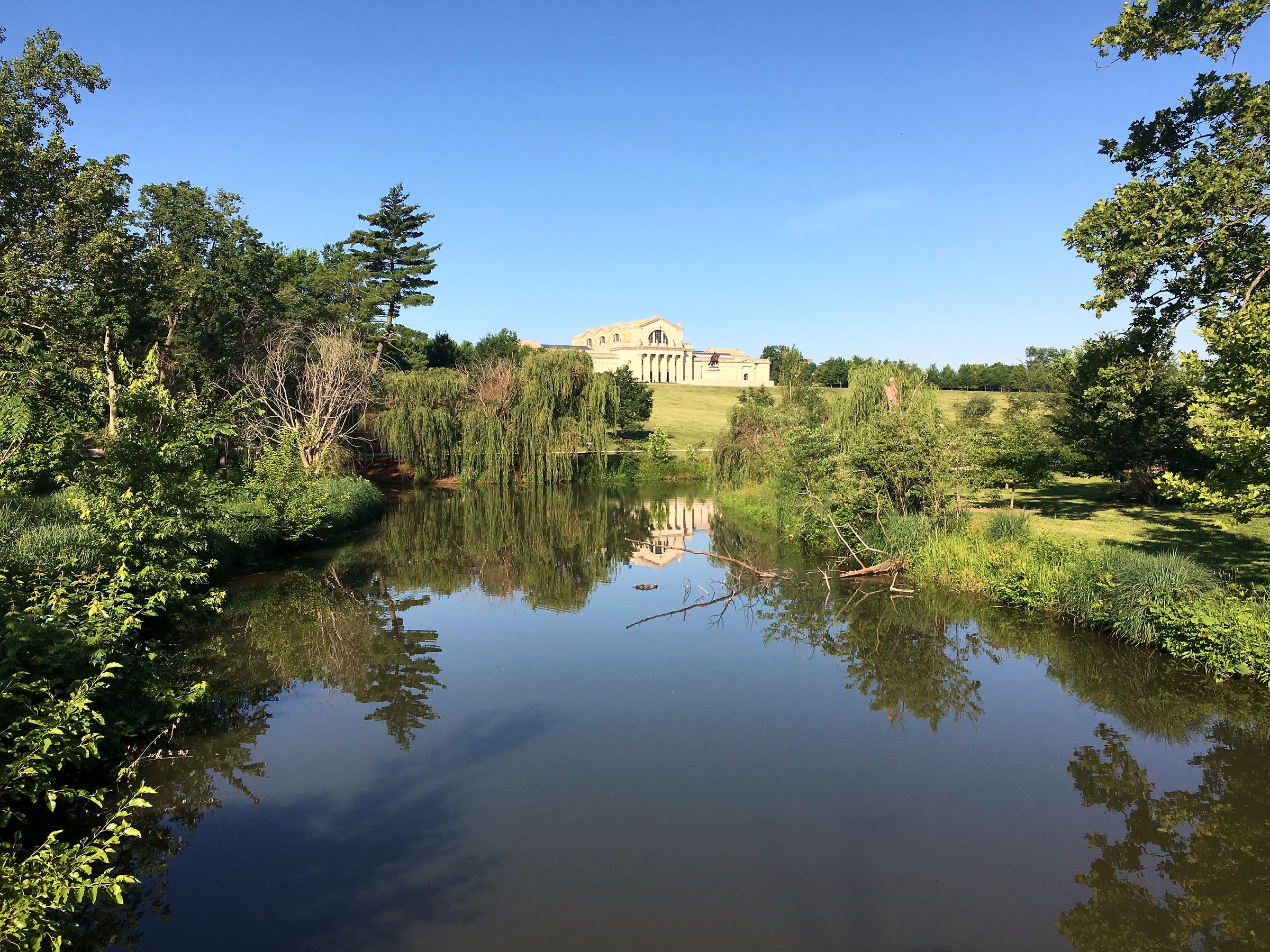 Calm pond mirroring willows and a stone portico on a green hill.