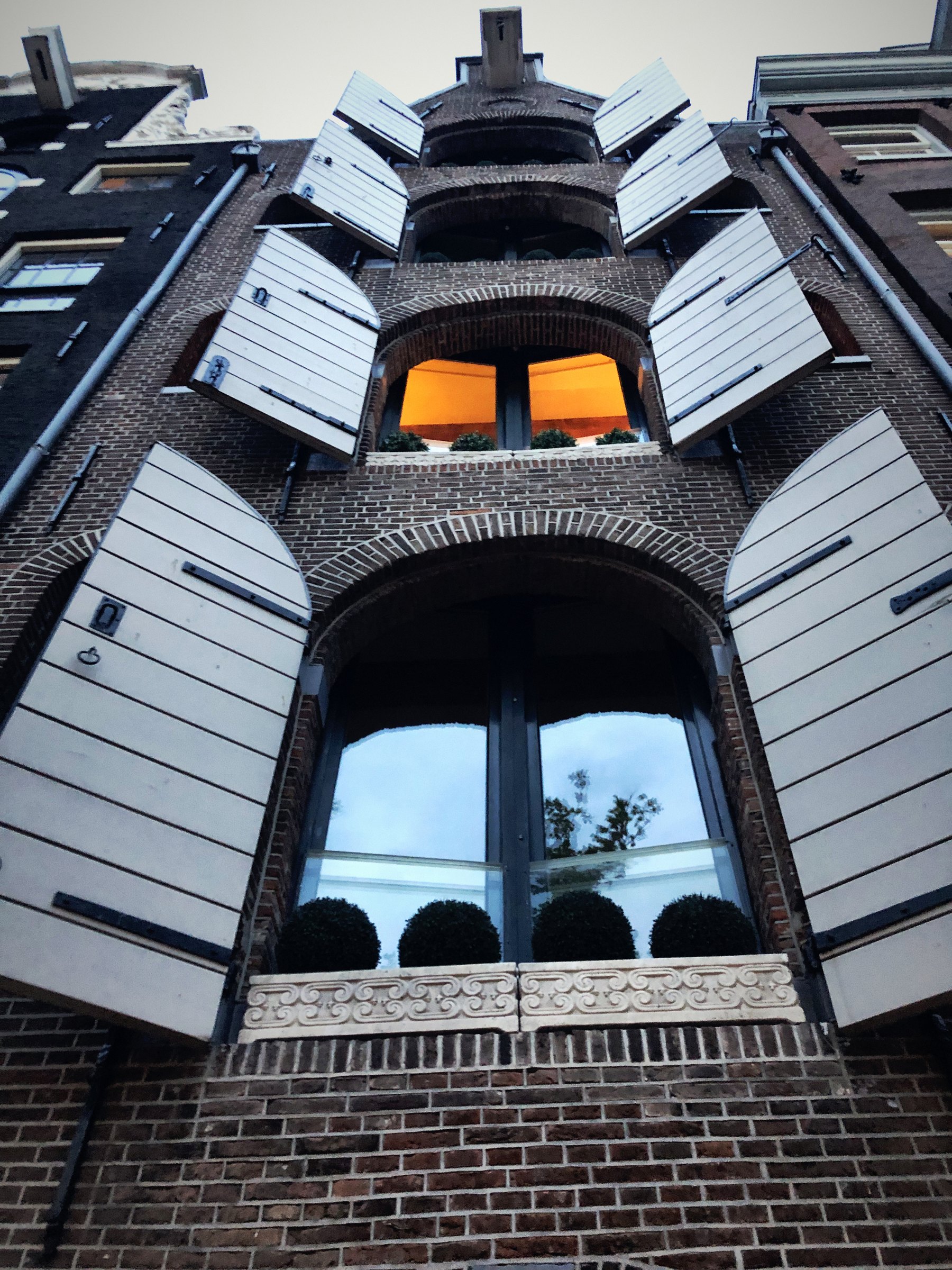 Looking straight up a facade of white wooden shutters around a lit window.