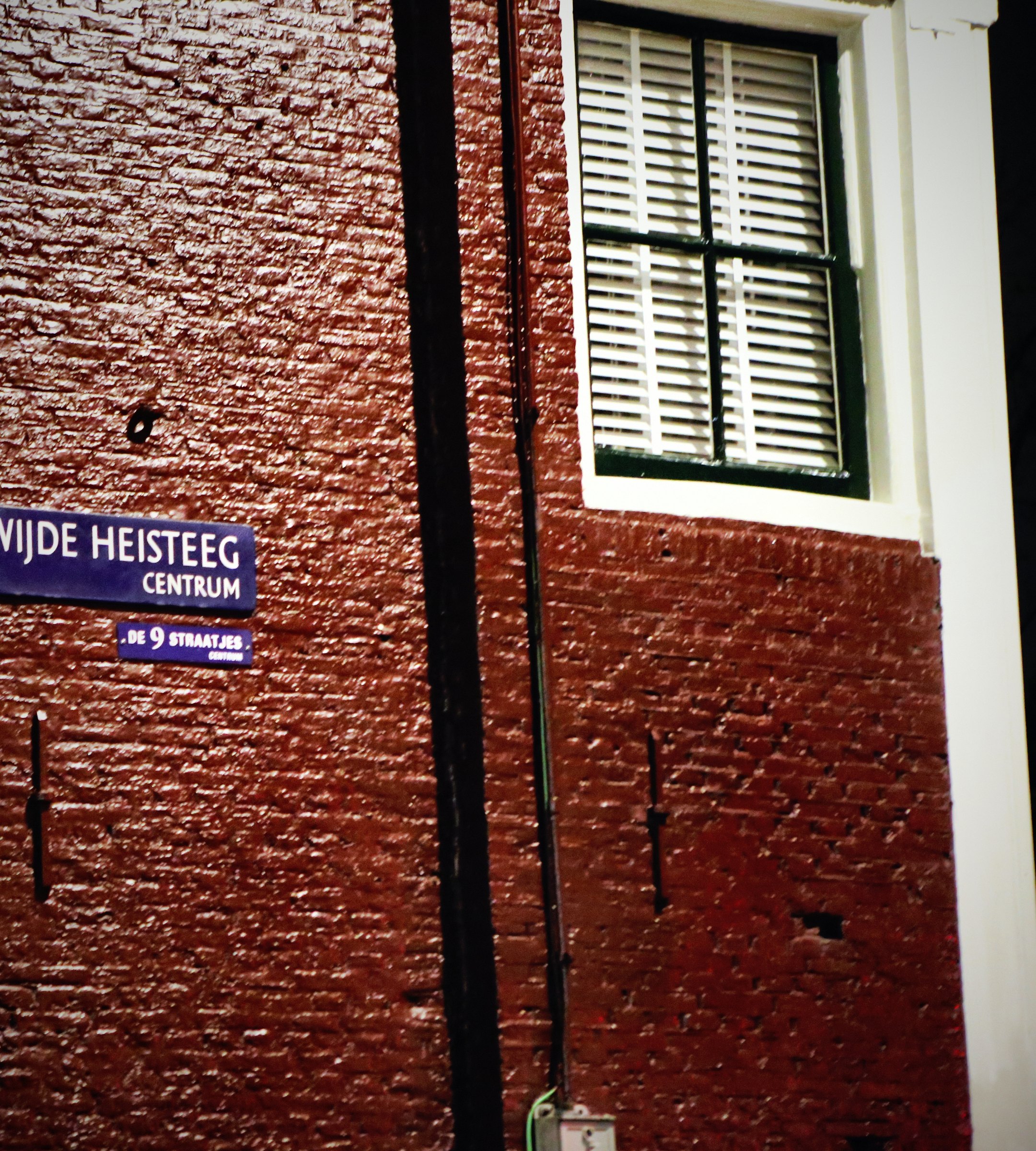 A red brick wall with a white-framed window and a black drainpipe beside a street sign.