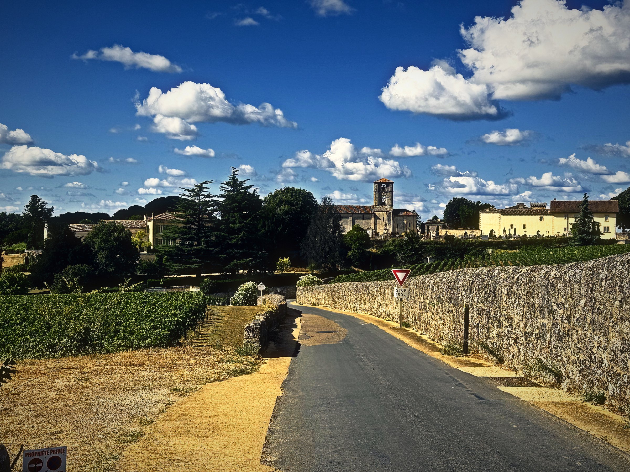 A country road running beside a long limestone wall toward a distant village and tower under scattered clouds.