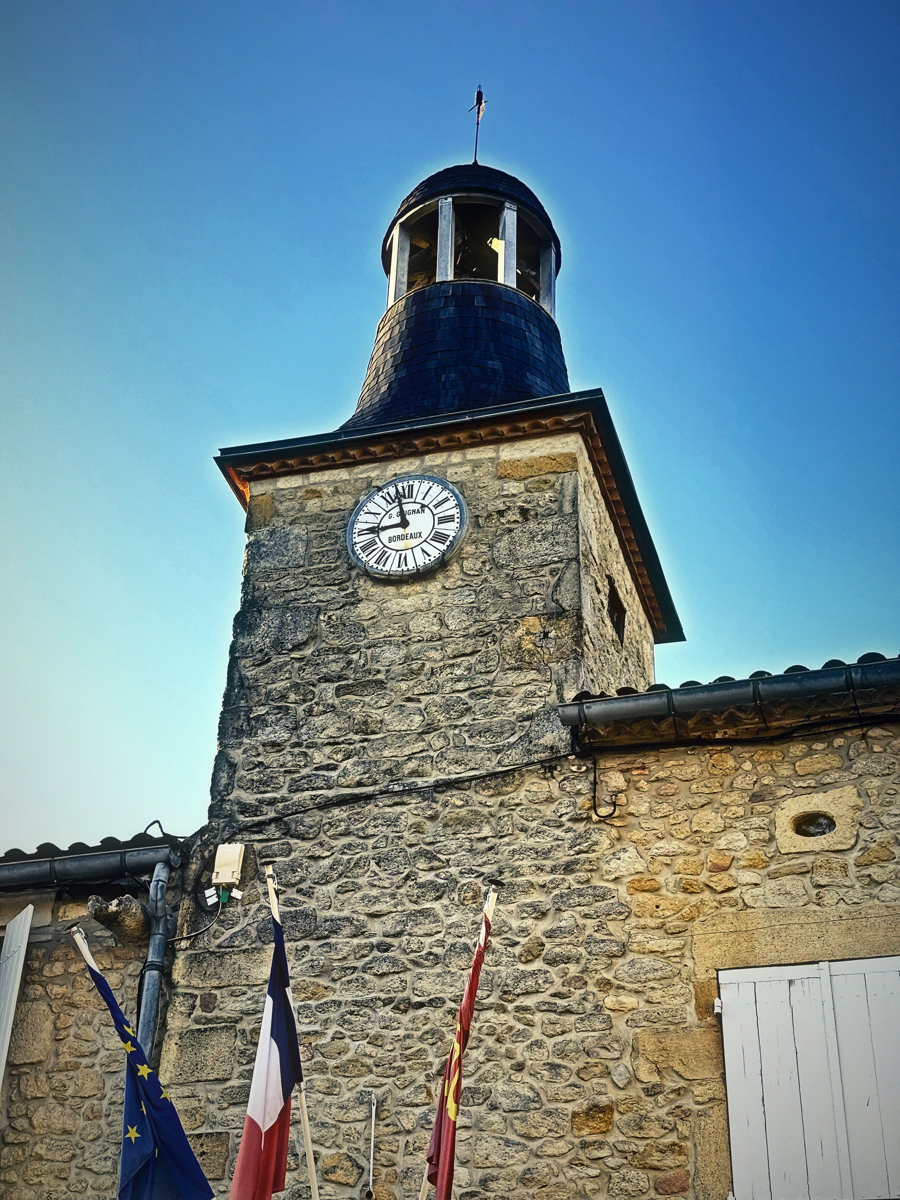 A stone clock tower with a slate bell cupola above a town hall flying French flags.