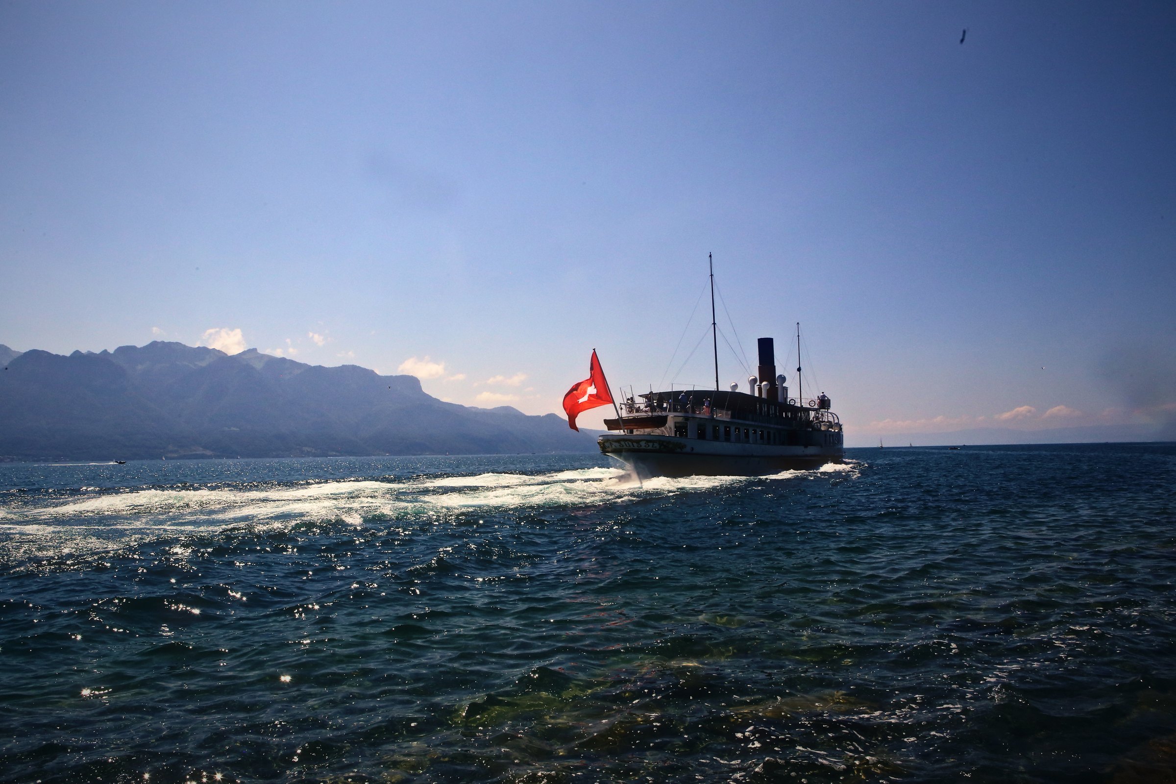 Paddle steamer with a red Swiss flag leaving a white wake on Lake Geneva, mountains behind.