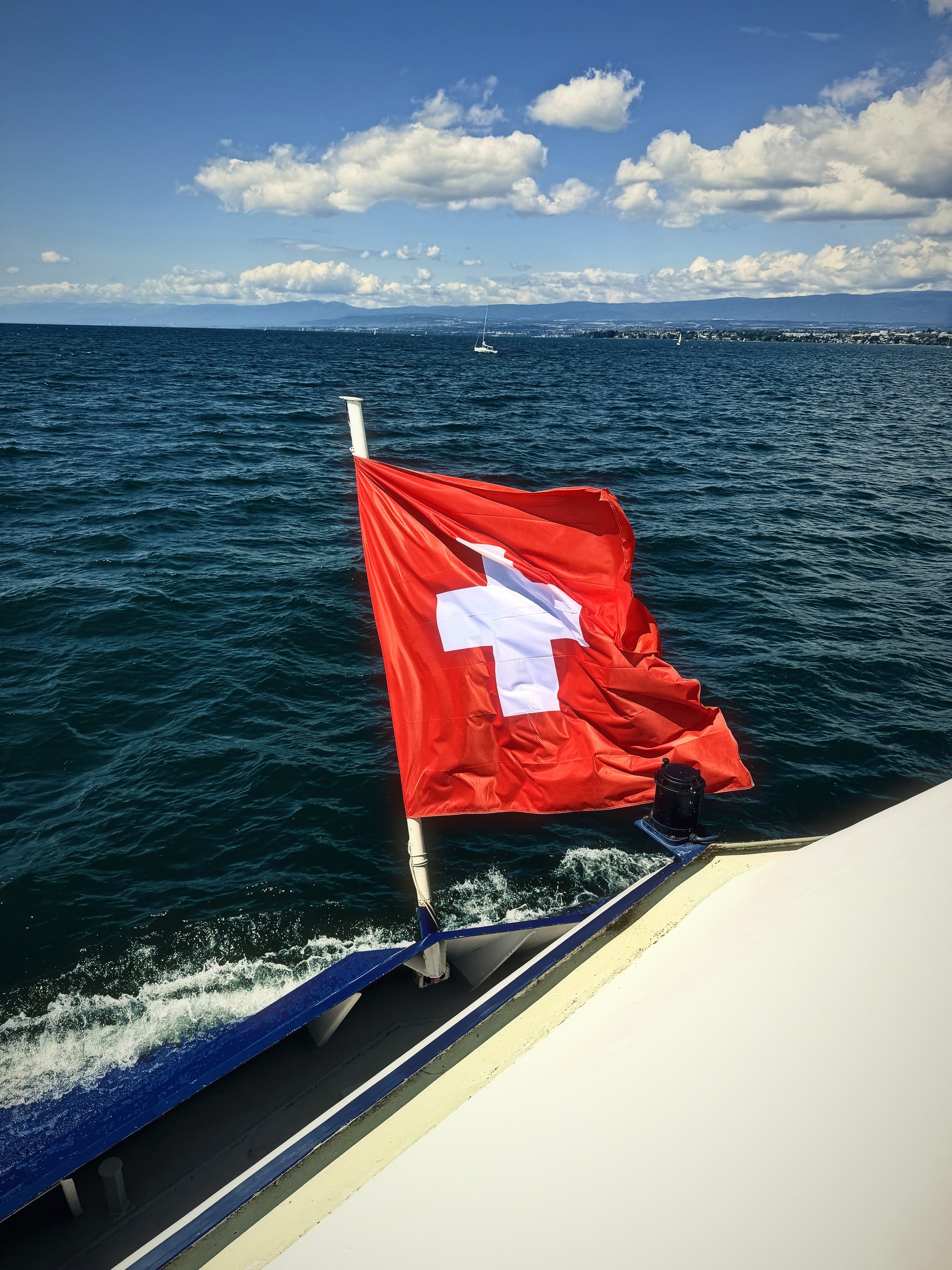 Red Swiss flag with a white cross flying over the side of a boat on blue water.