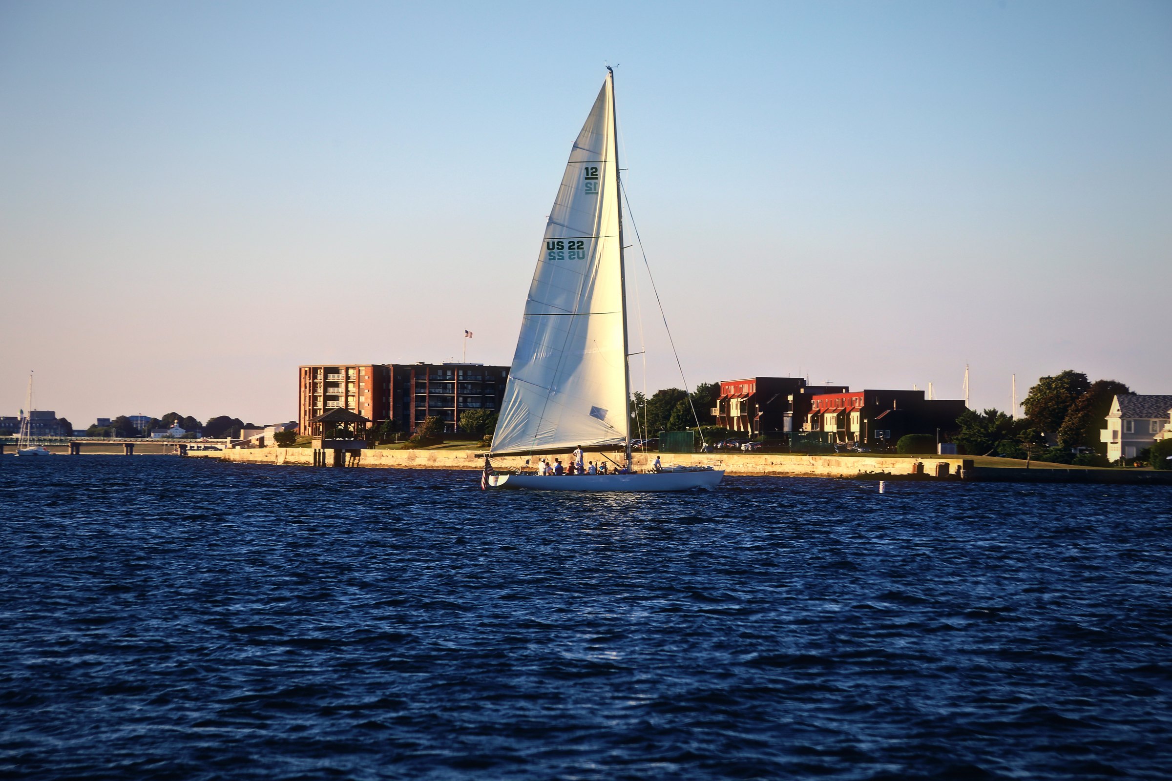 White sail crossing a harbor in front of a low brick waterfront building.