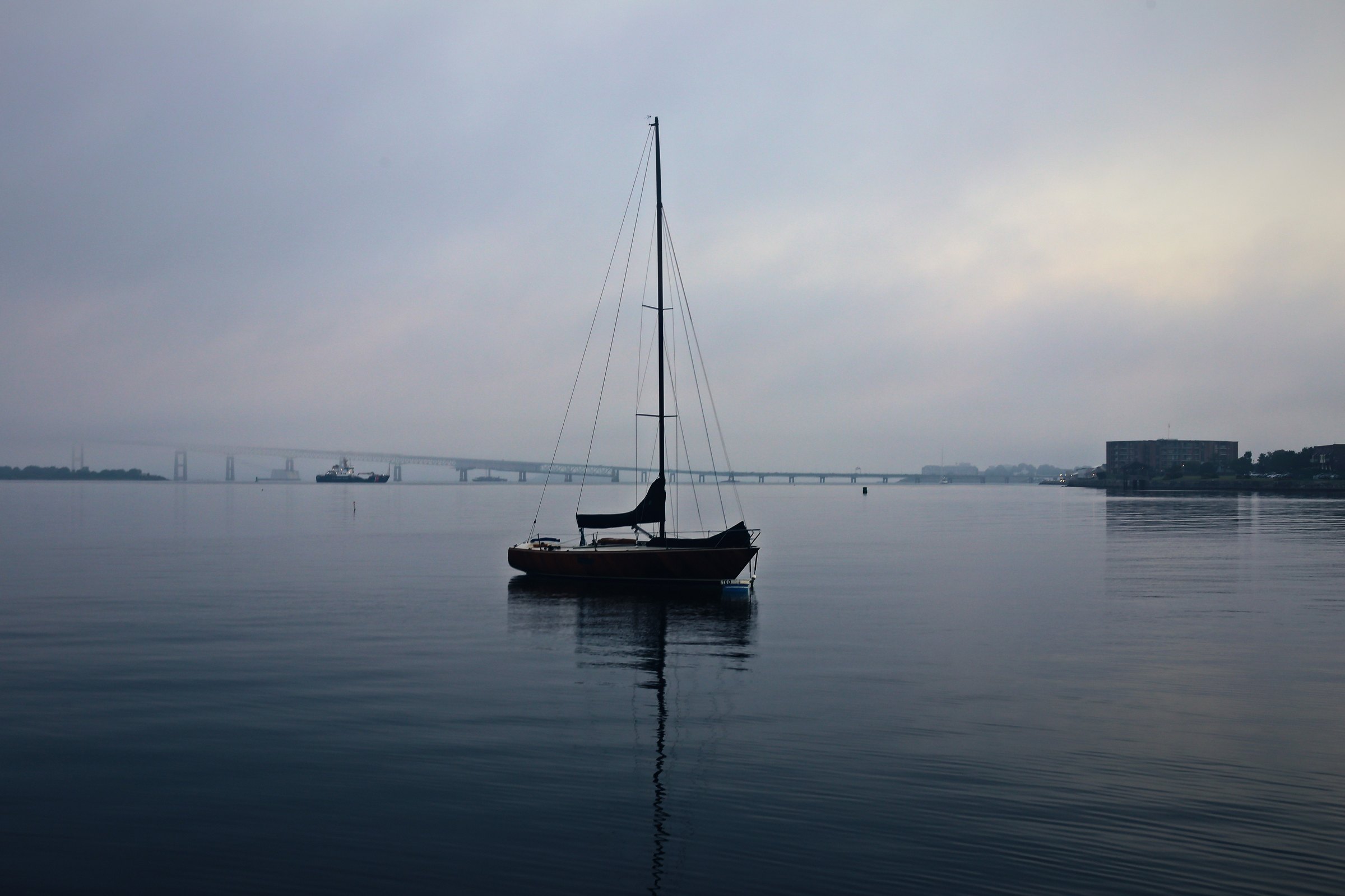 Lone sailboat on glassy, misty grey water with a faint shoreline.
