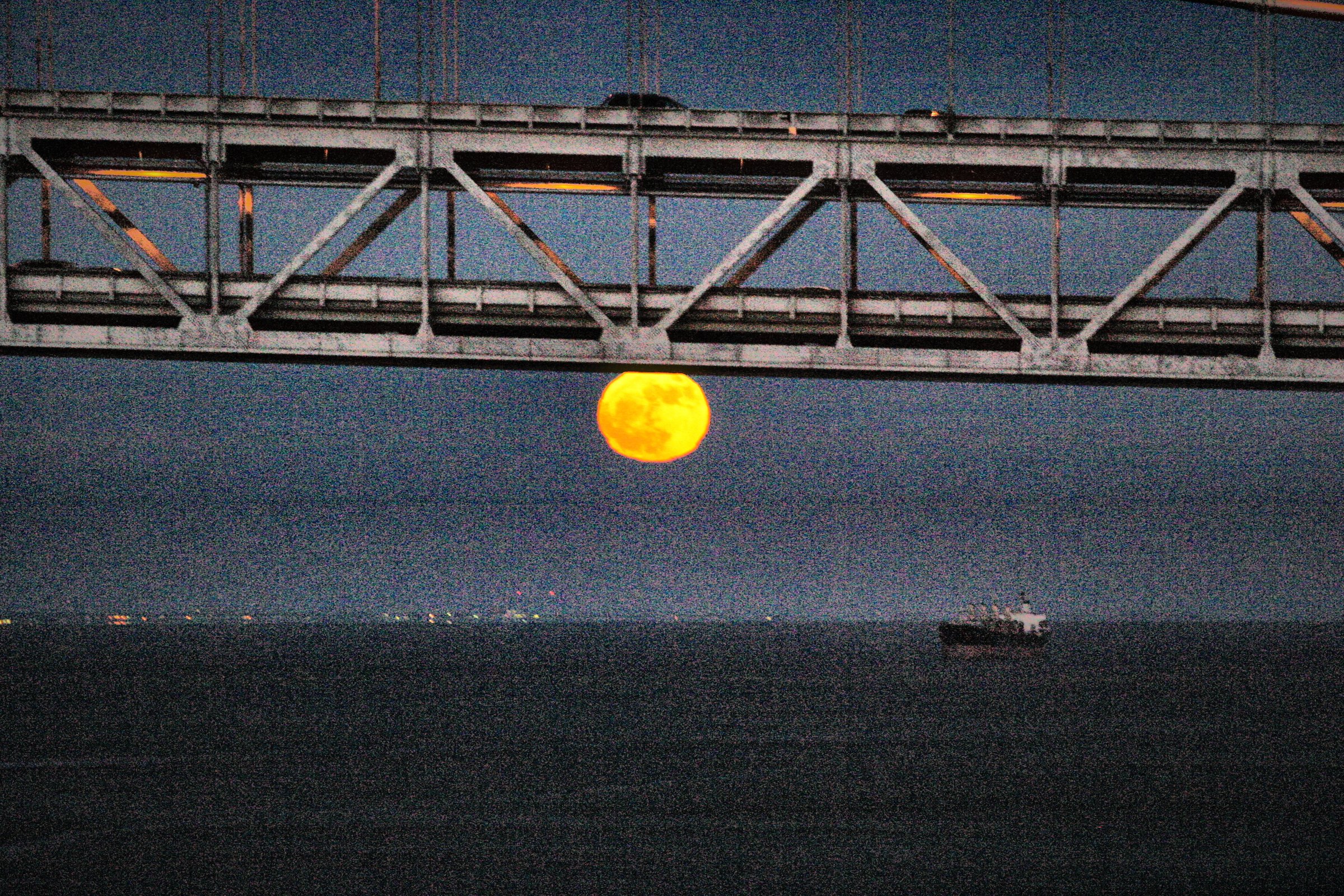 A huge orange full moon rising beneath the dark steel truss of a bridge, a few ships far off on the water.