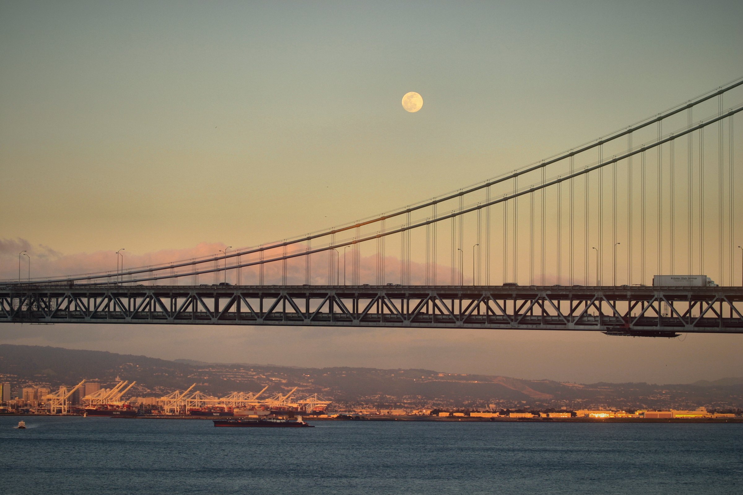 A small full moon climbing above a long suspension bridge across a bay in warm dusk light.