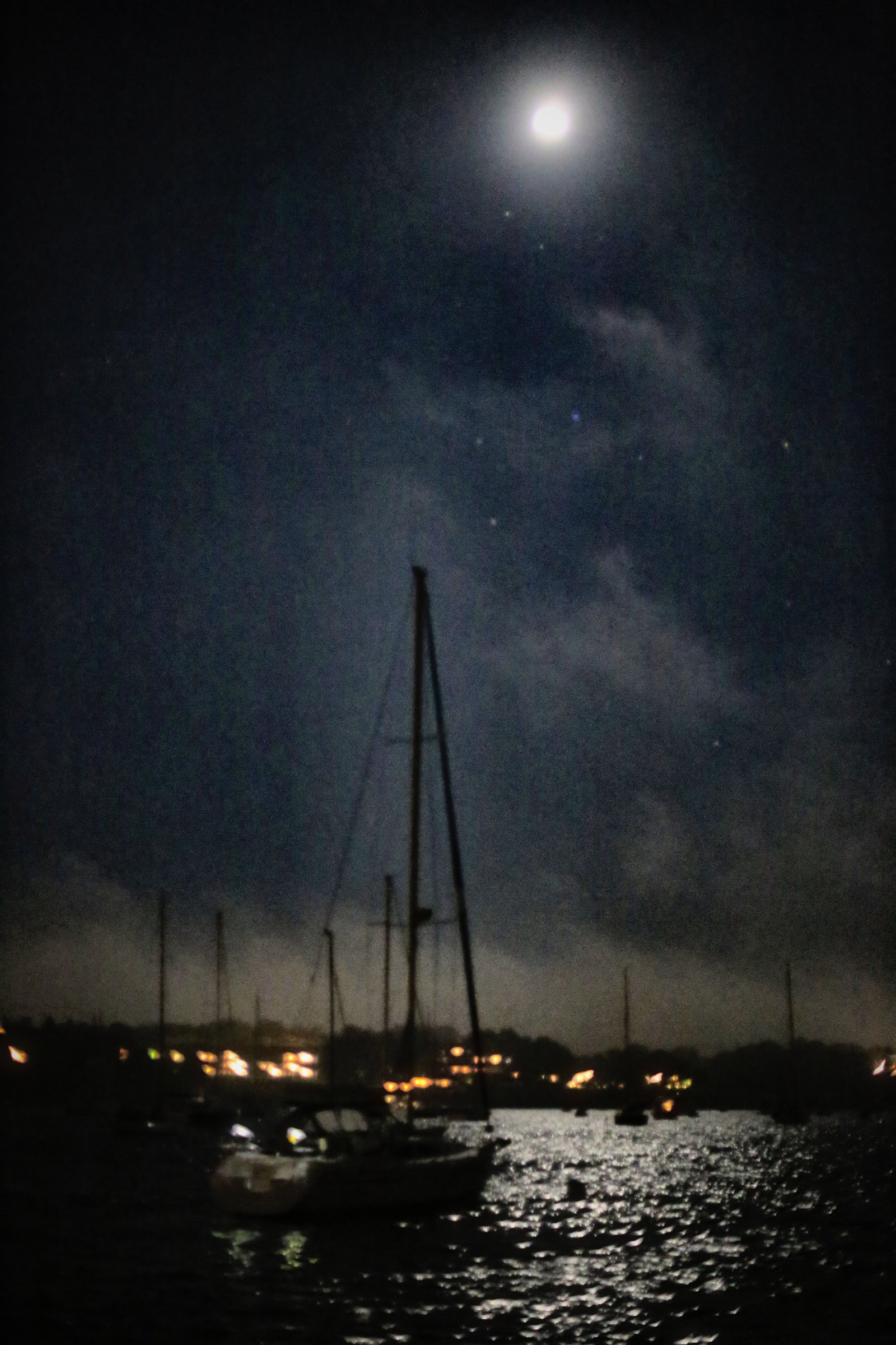 A bright moon high over a harbour at night, a sailboat mast reaching up toward it and lights shimmering on the water.