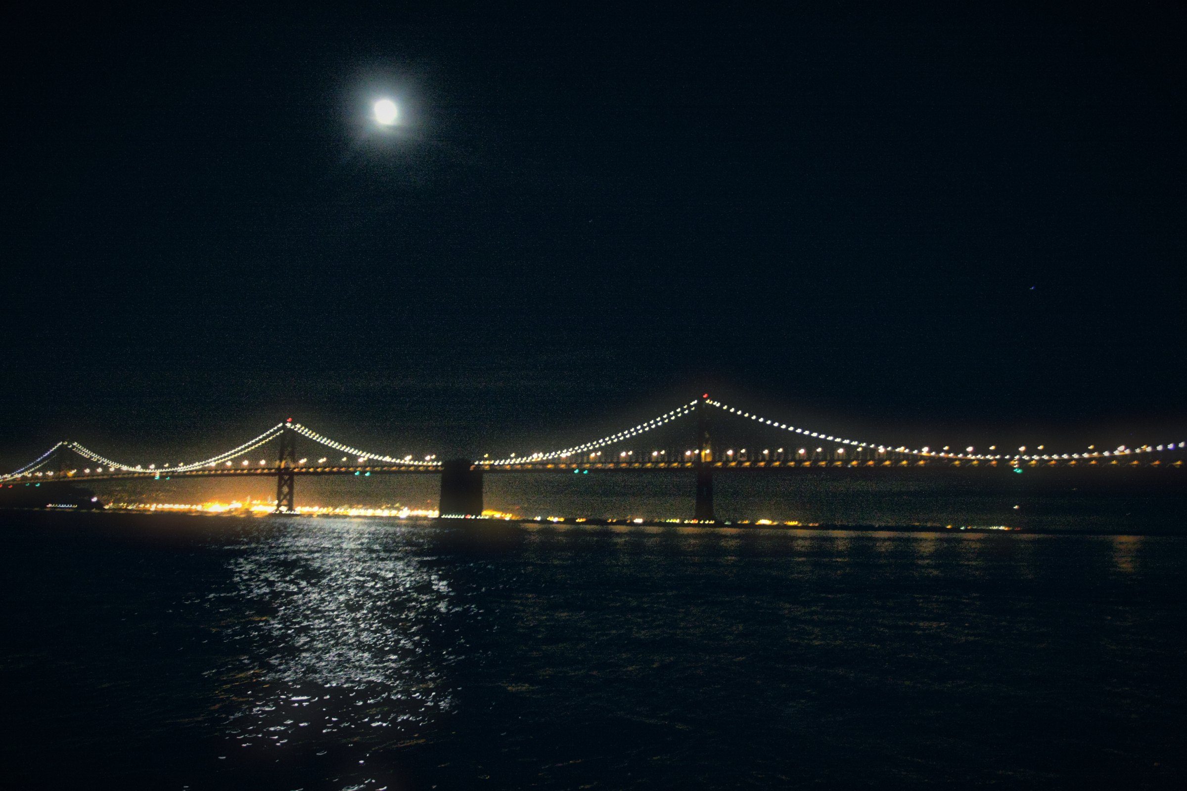 A bright moon above the dark bay, the Bay Bridge strung with glowing lights and their reflections across the water.