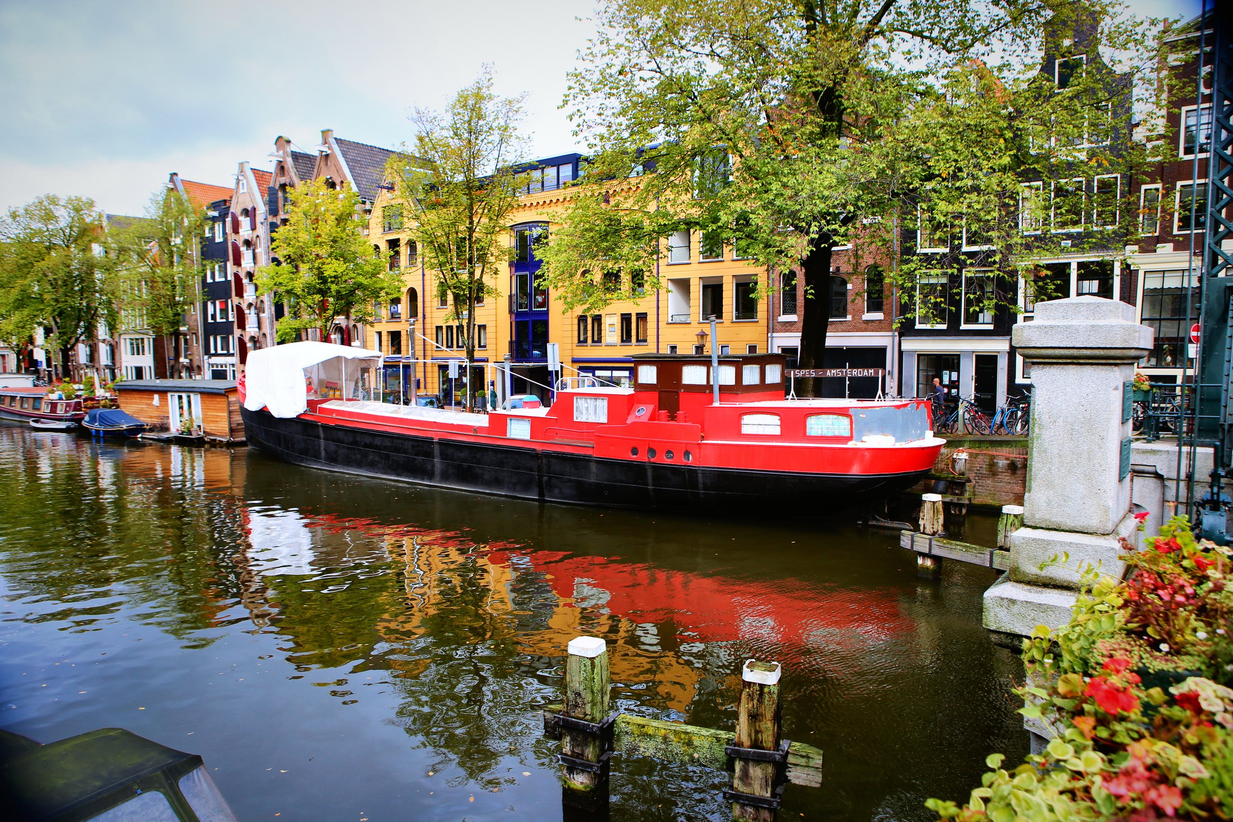 A red houseboat on a canal reflecting gabled houses, red flowers in the corner.