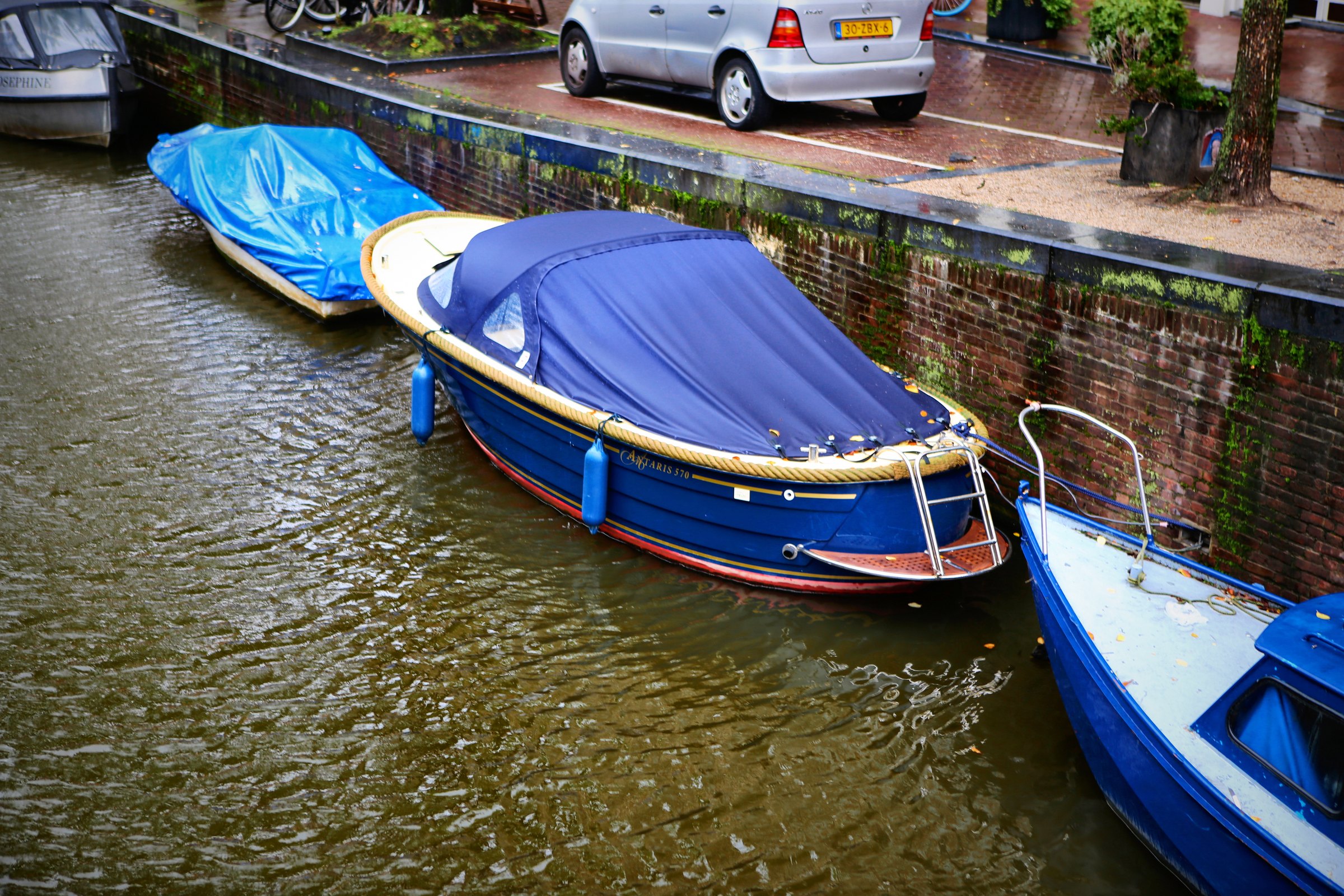Boats under bright blue covers moored along a brick canal wall.