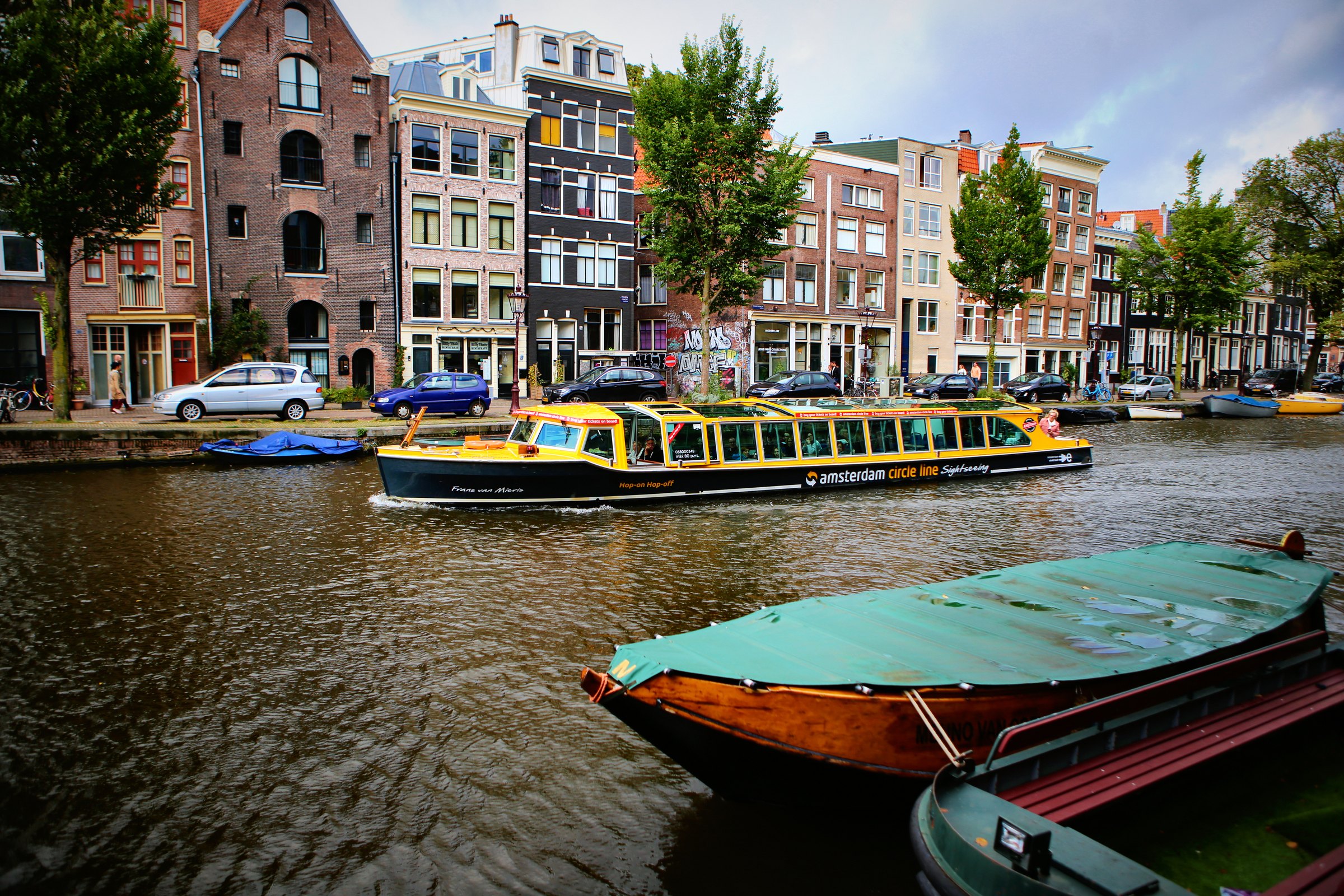 Covered boats in green and red moored on a canal, a yellow canal boat passing gabled houses behind.