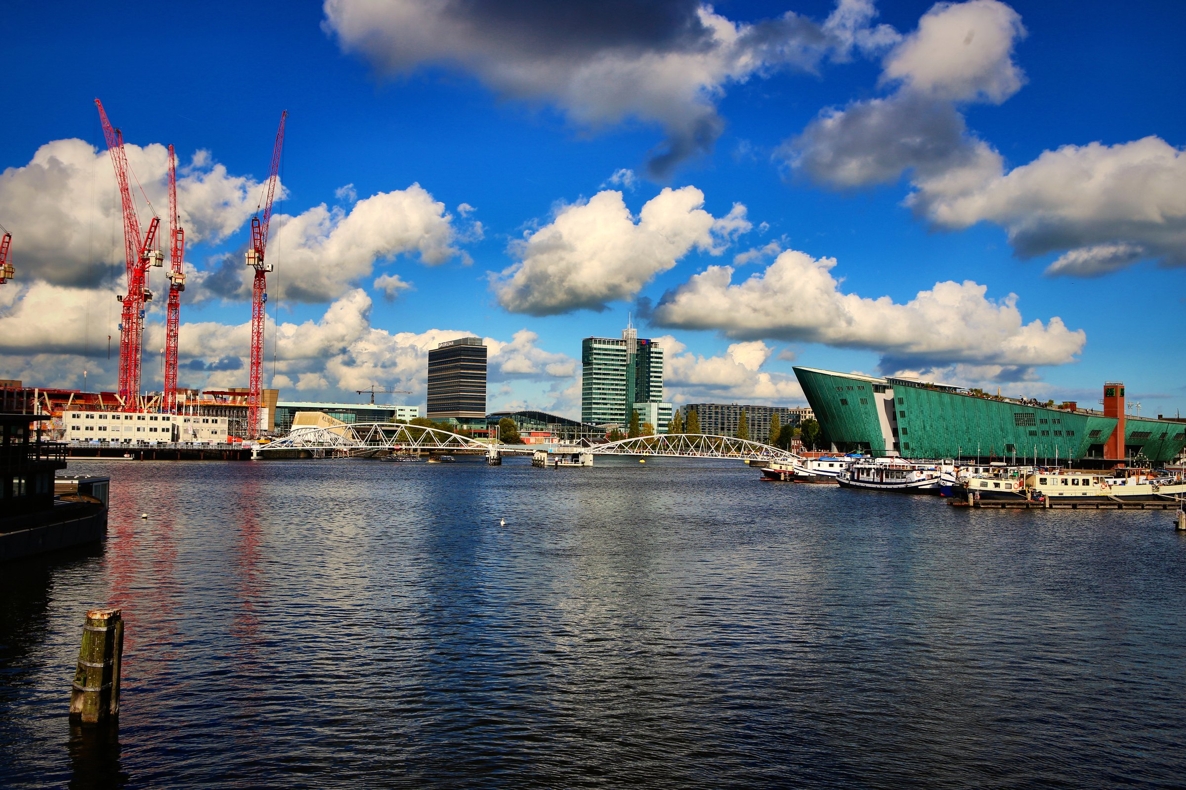 Red harbour cranes and a green ship-shaped building across open water under big clouds.