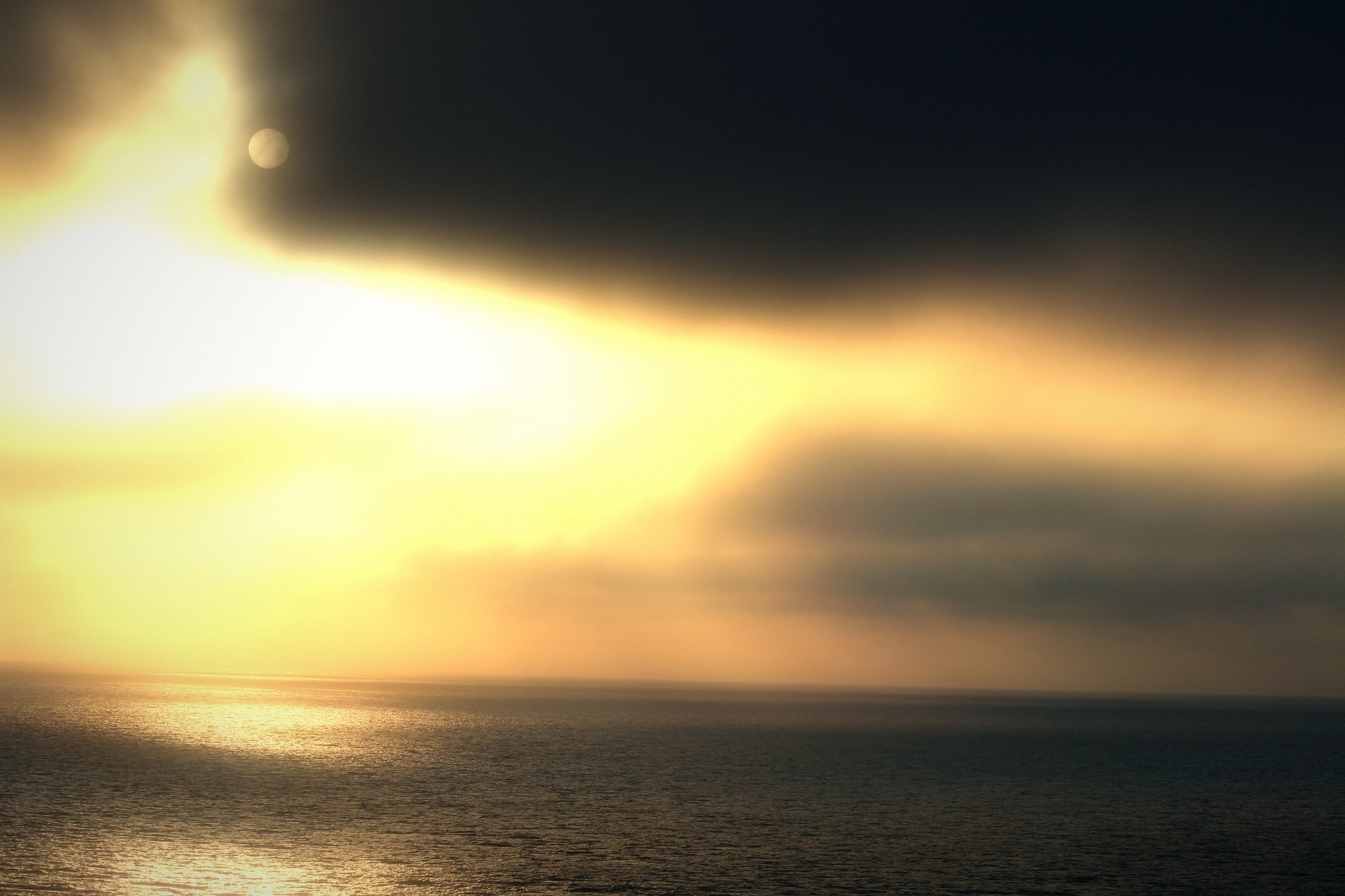 Glowing golden light behind dark clouds over a bright, calm sea.