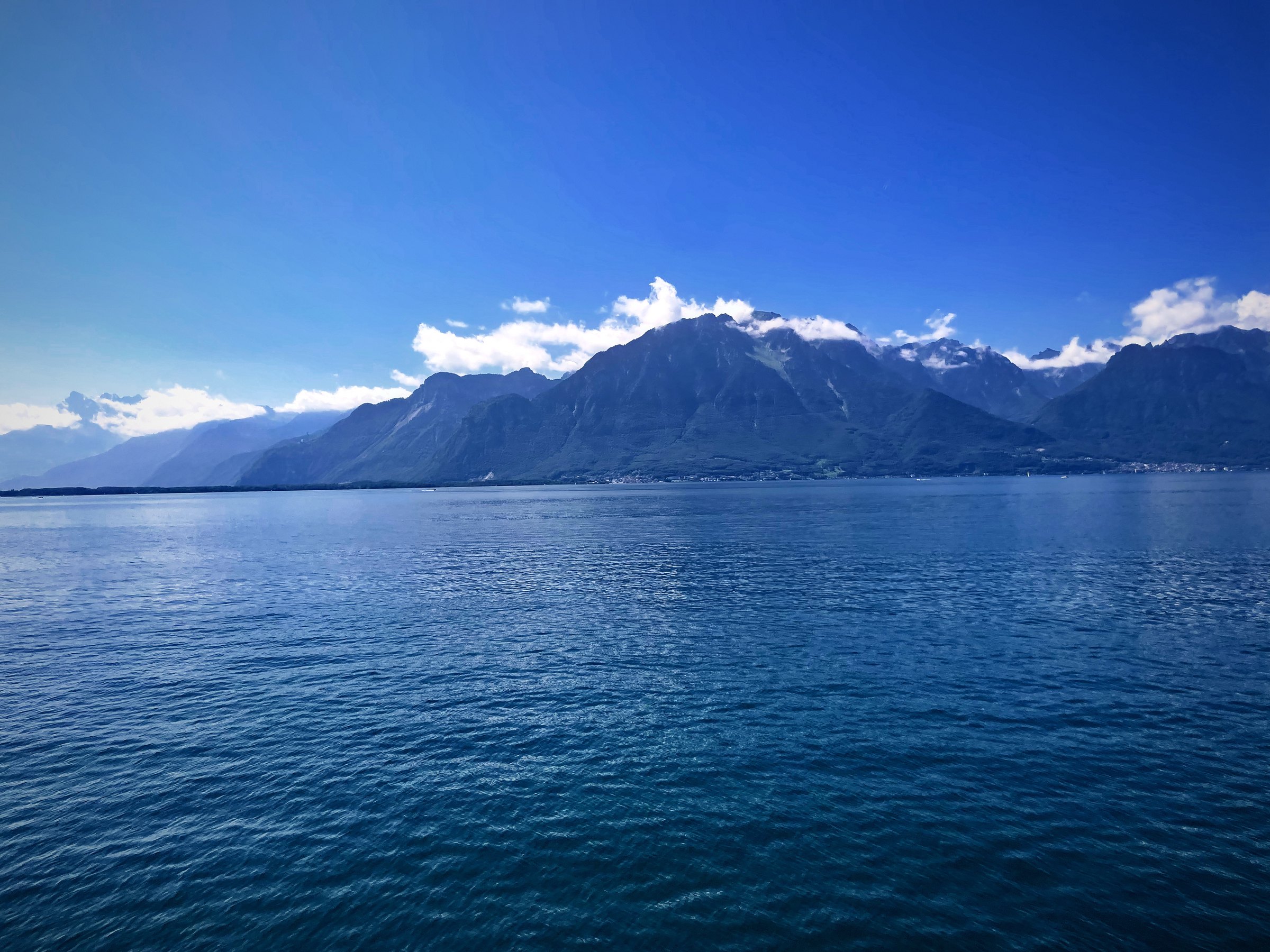 Deep teal Lake Geneva water stretching to snow-streaked peaks under bright clouds.