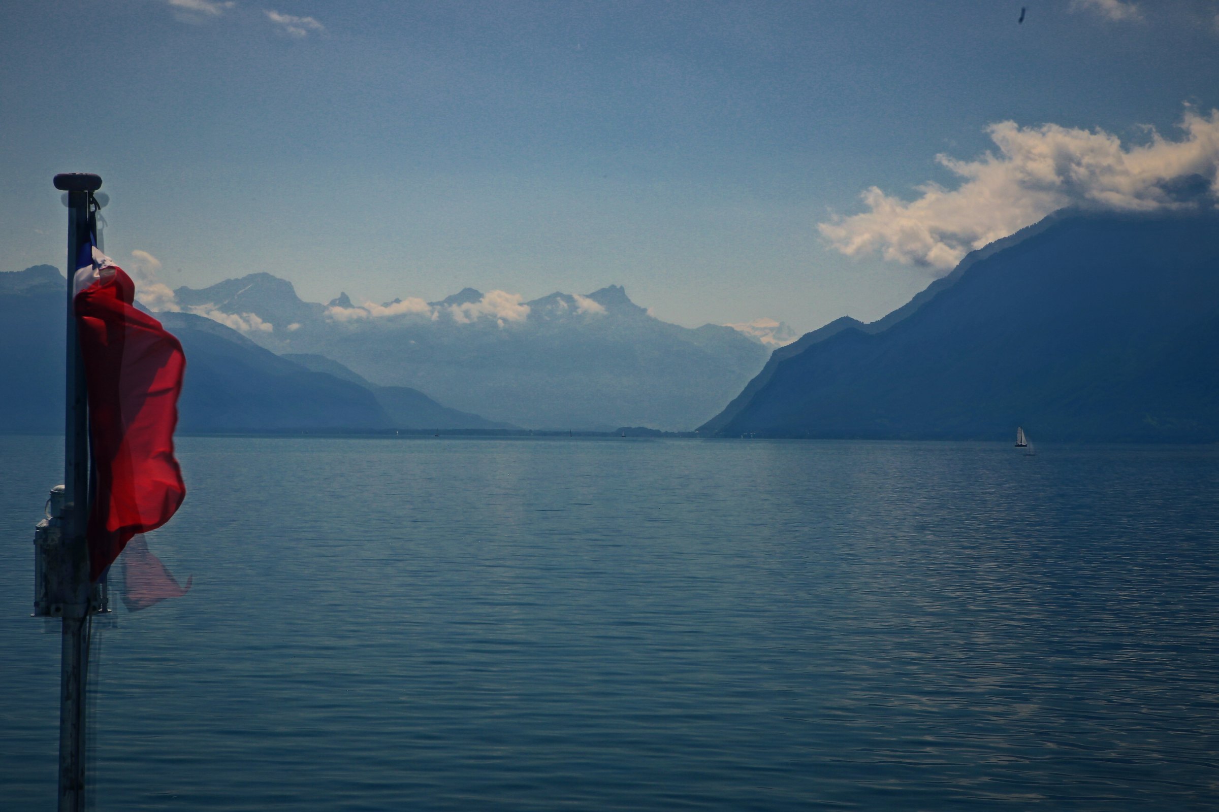 A red flag on a post beside calm blue lake water with mountains beyond.