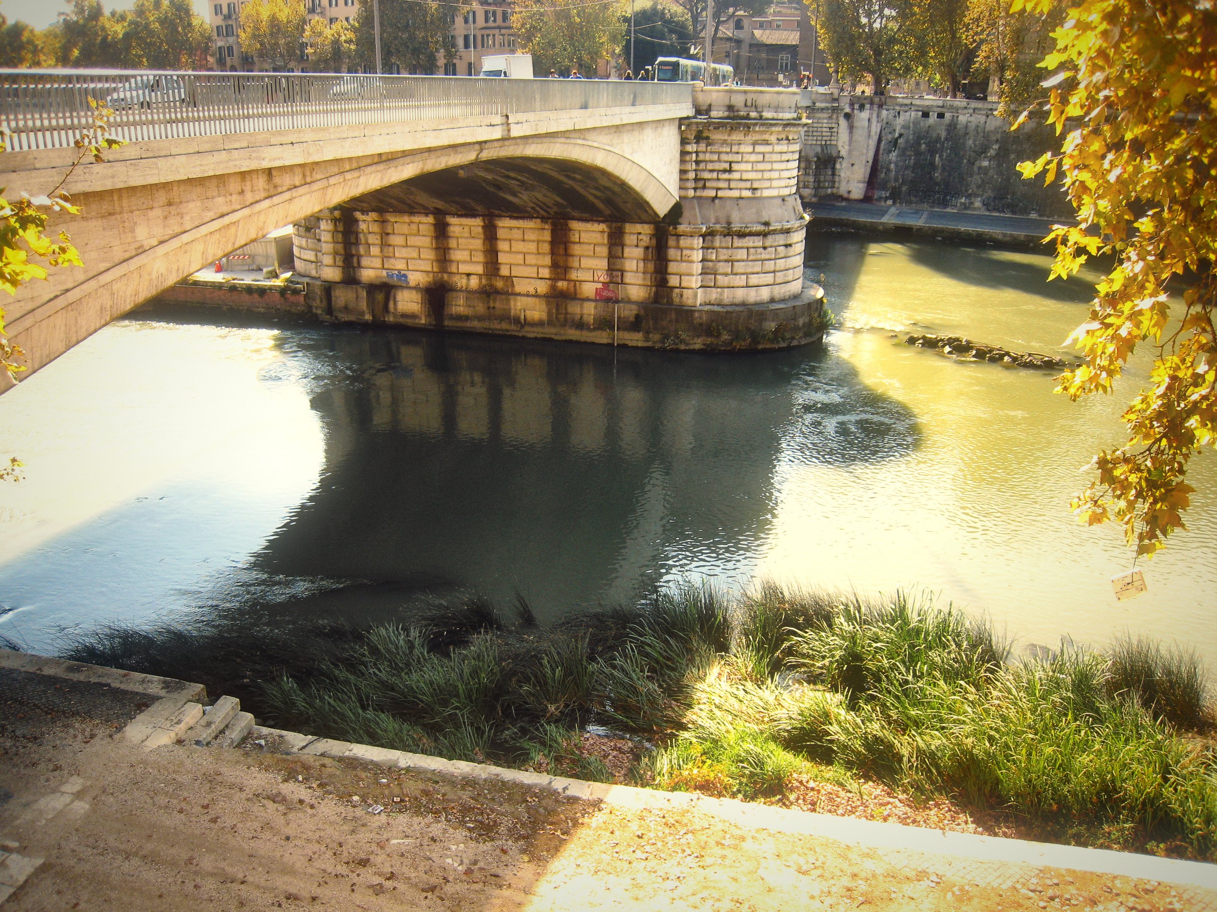 A bridge over the Tiber with sunlight on the water and the bank below.
