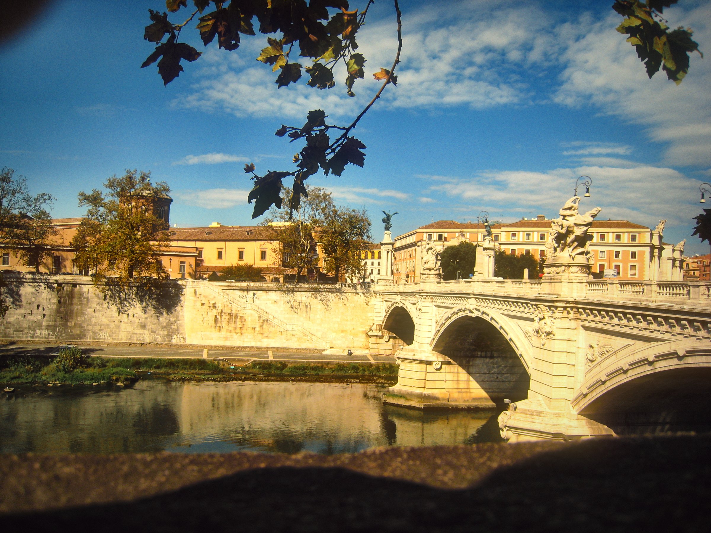 A stone bridge with statues over the Tiber, branches overhead.