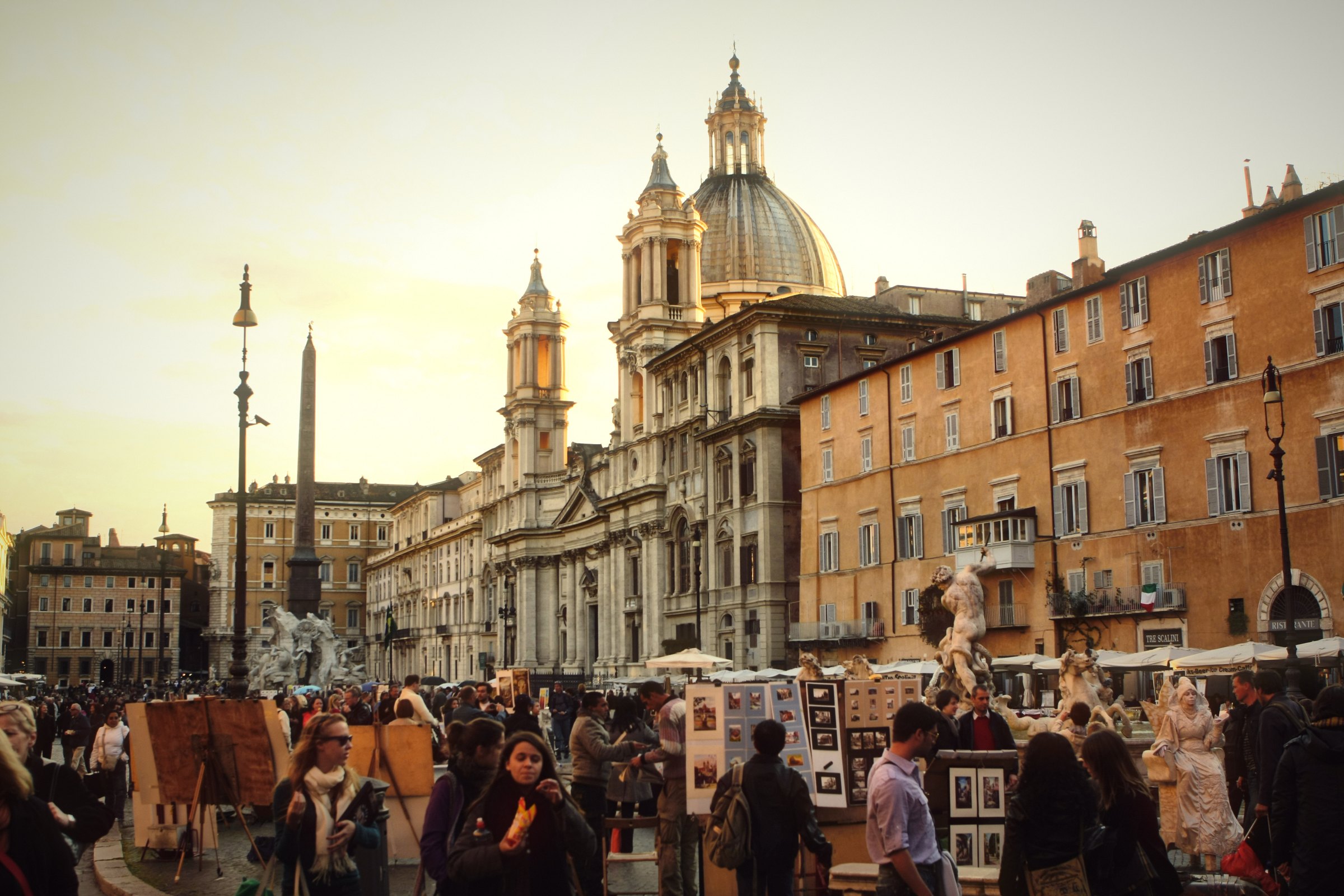 A crowded Piazza Navona with church domes in soft gold light.