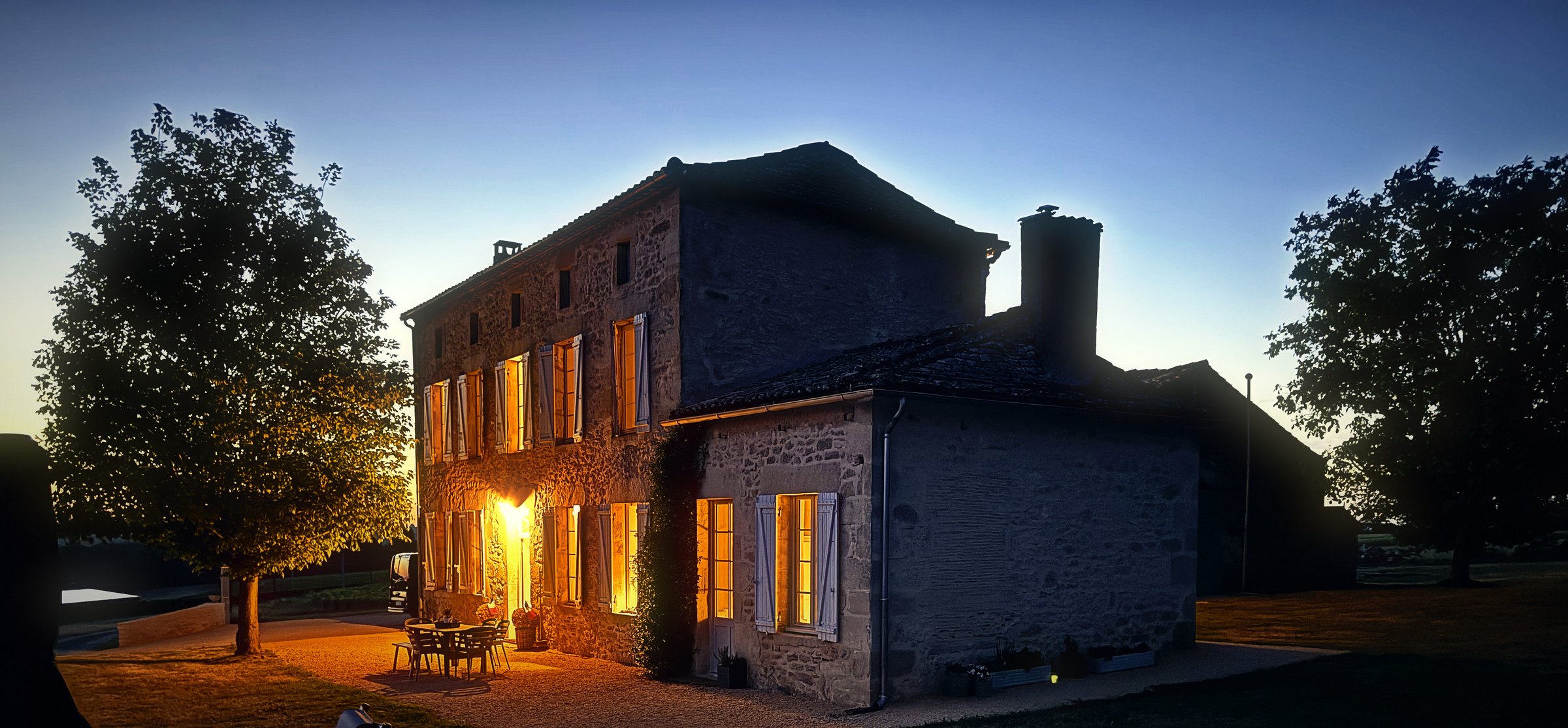 A stone house glowing with a lit lantern at dusk, framed by trees under a deepening sky.