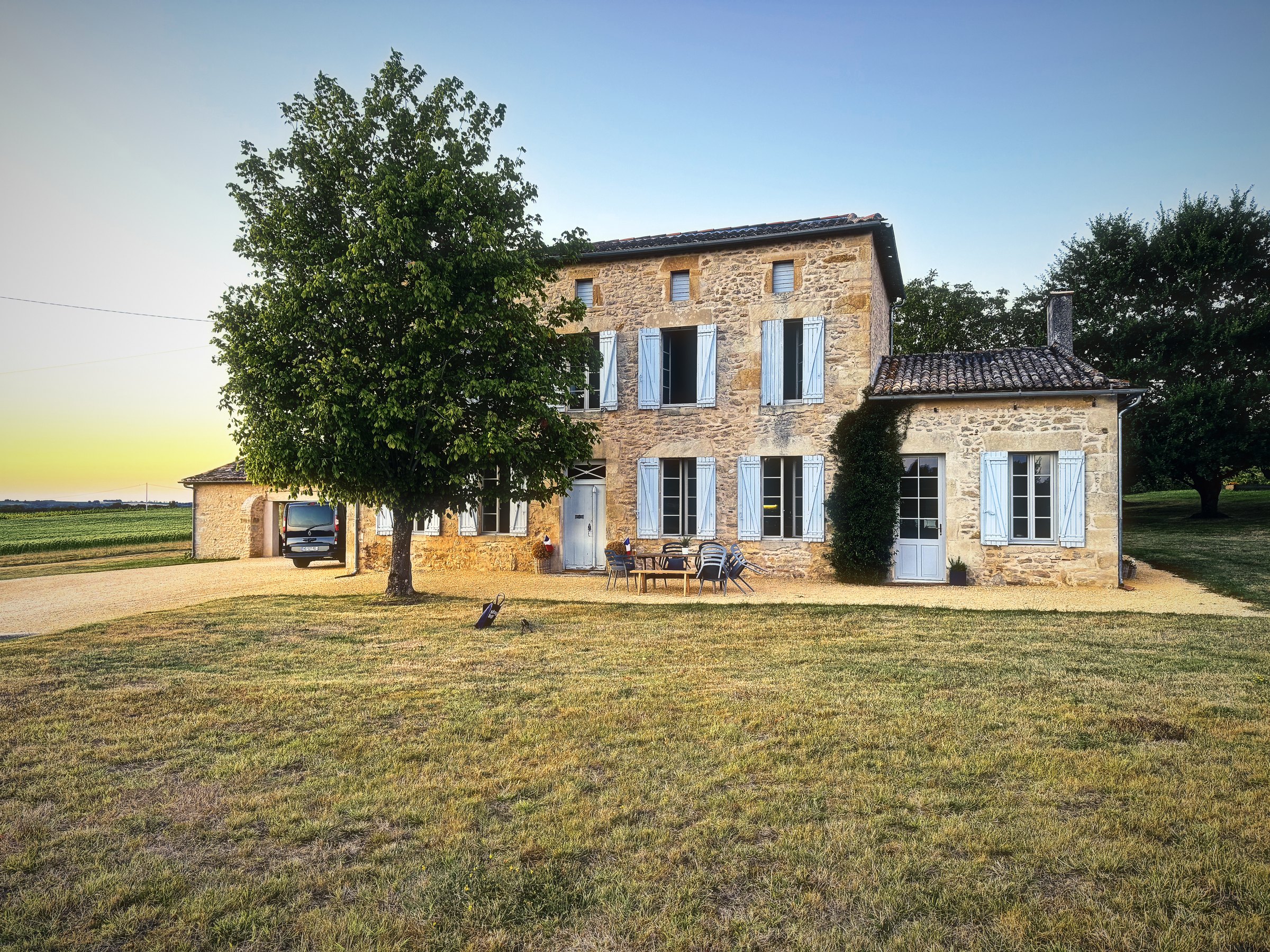 A stone farmhouse with blue shutters and a big tree, lit warm by the low evening sun.