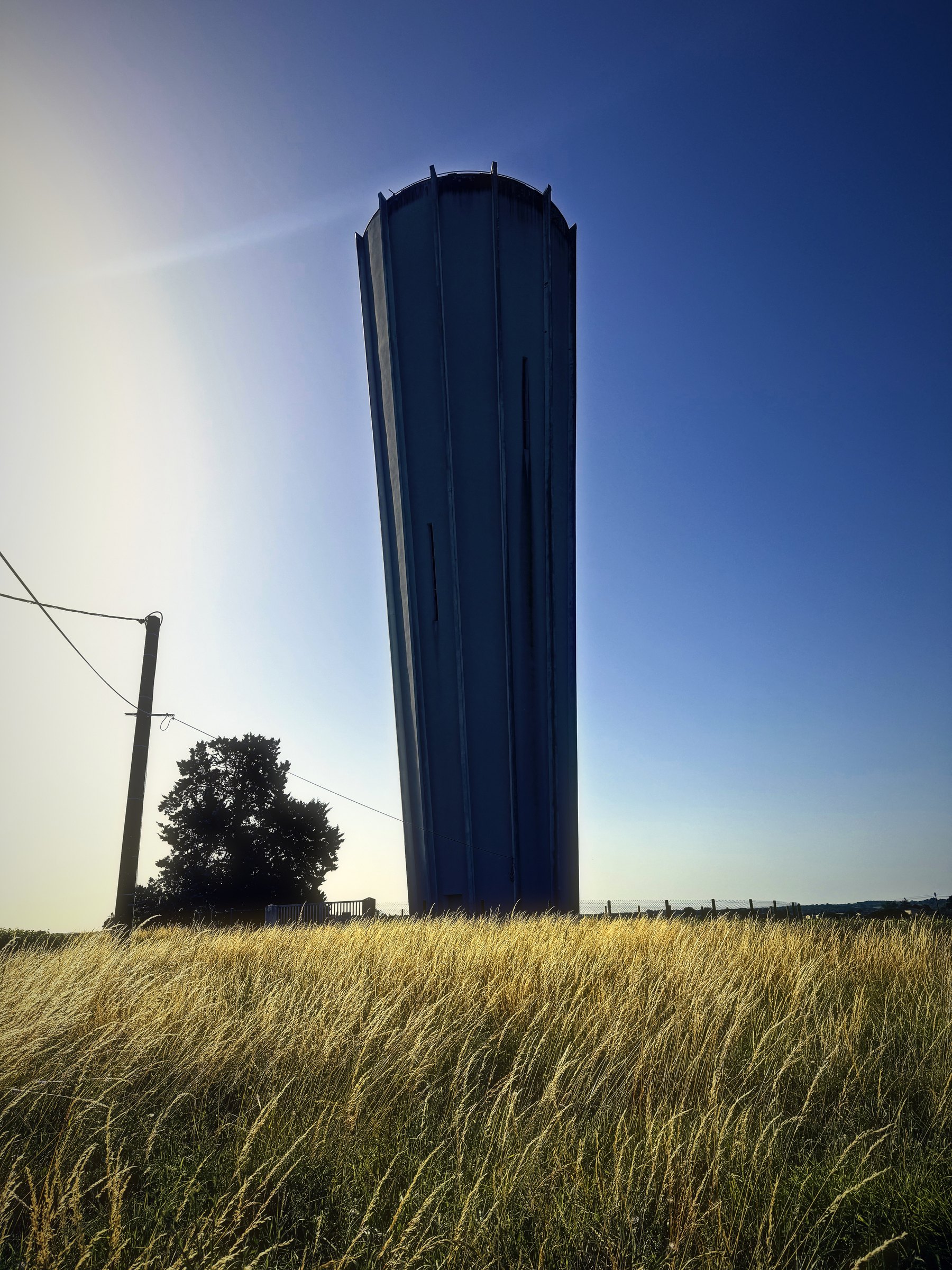 The tall concrete water tower above Pellegrue standing over gold summer grass as the morning sun flares beside it.