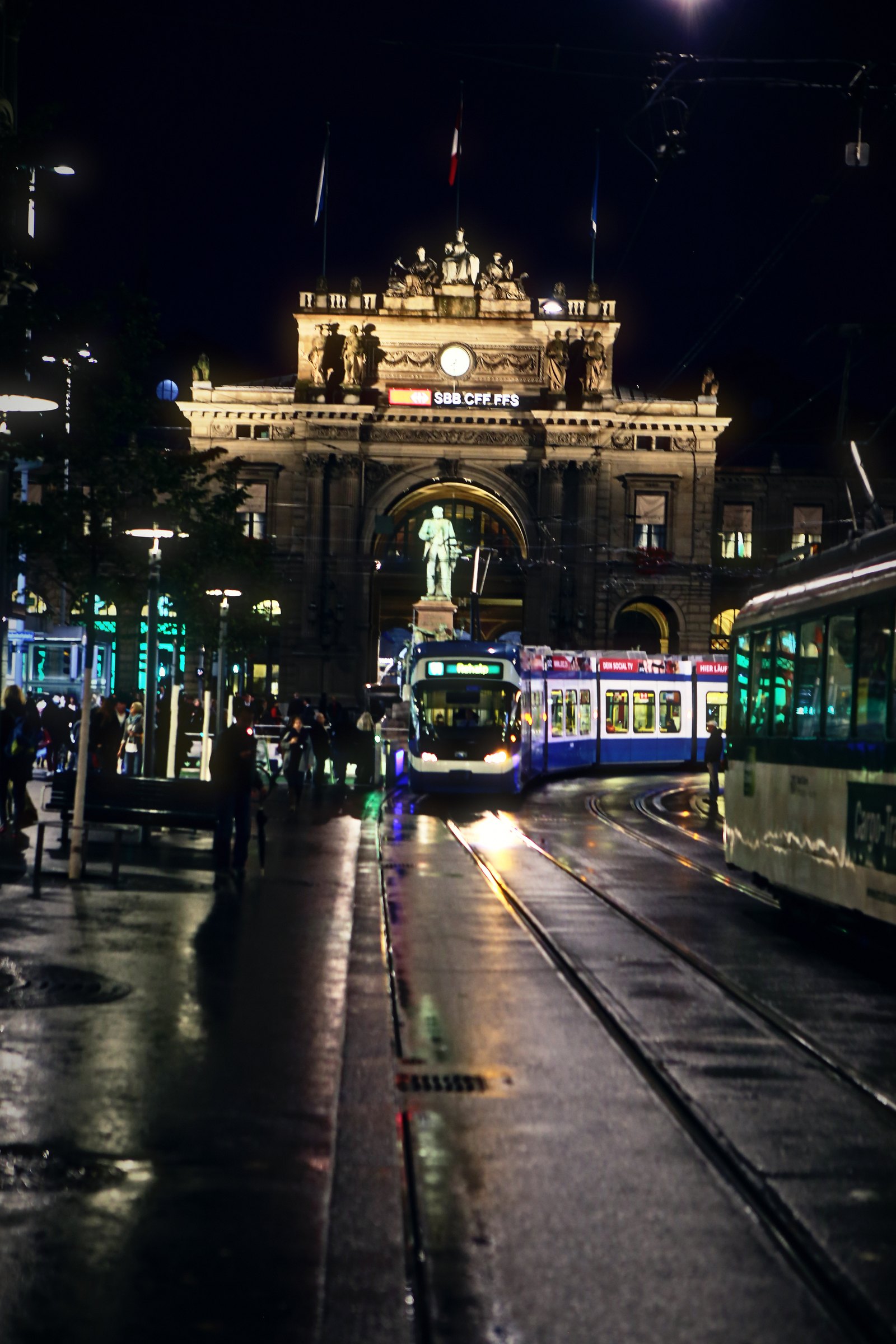 Floodlit stone station façade at night with a statue and wet tram tracks in front.