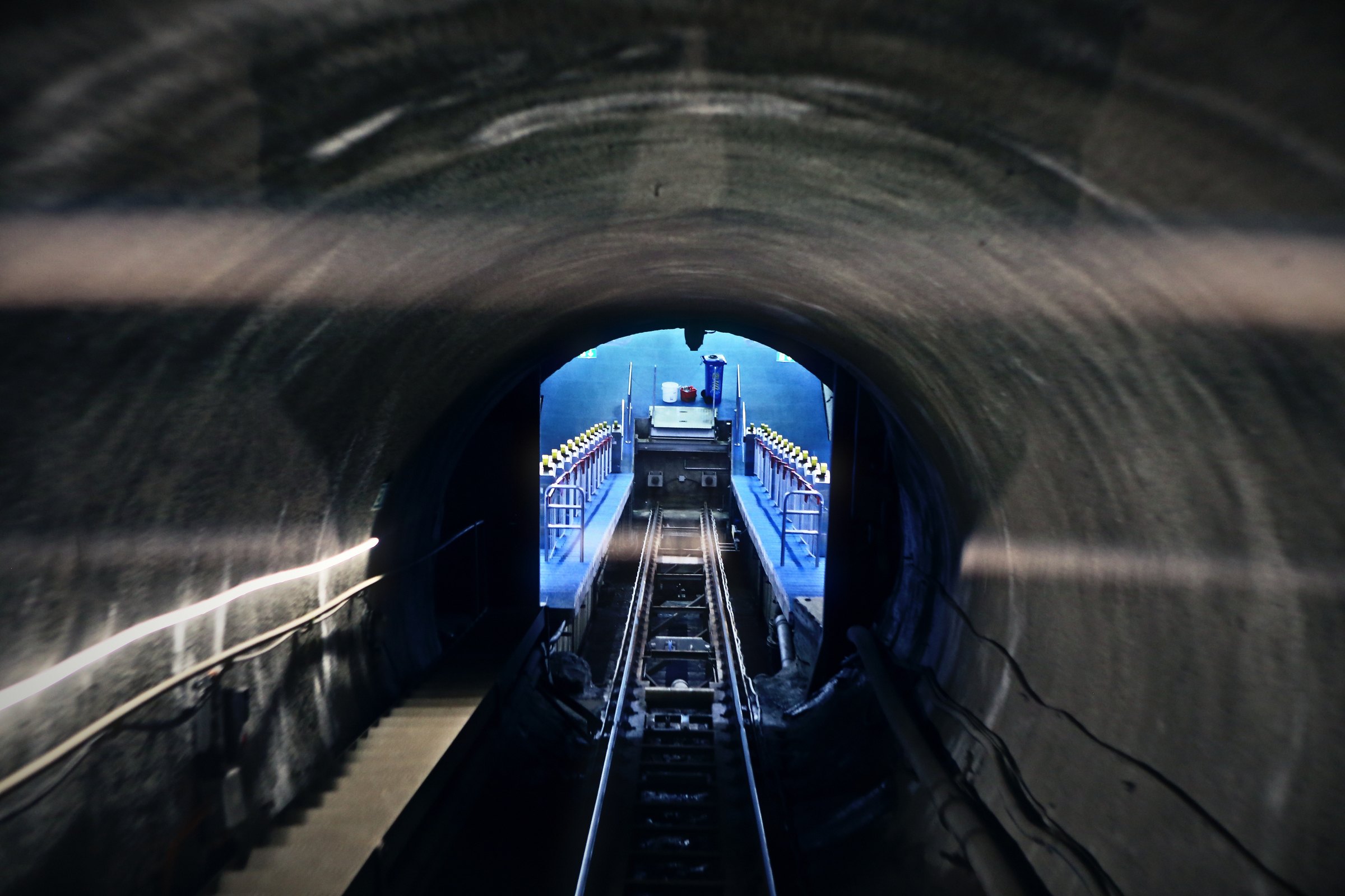 Round tunnel lined in rock with a lit funicular track vanishing at its center.