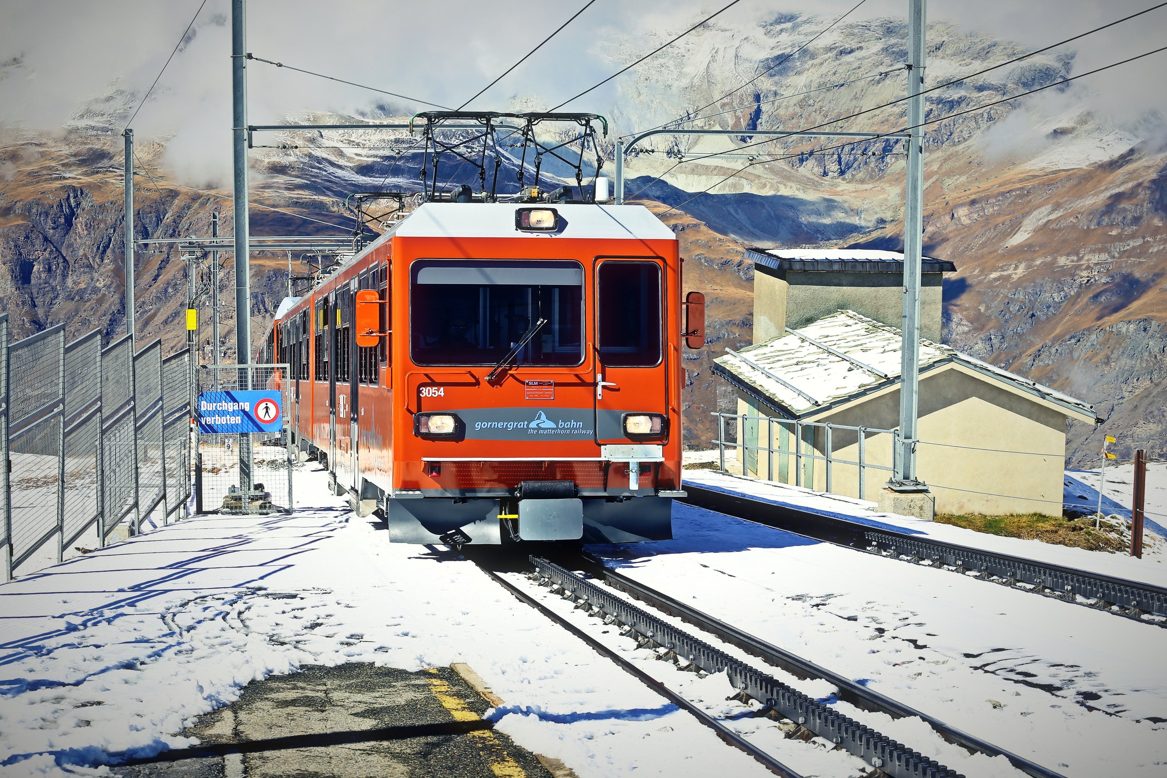 Orange mountain train at a snowy station with peaks behind.