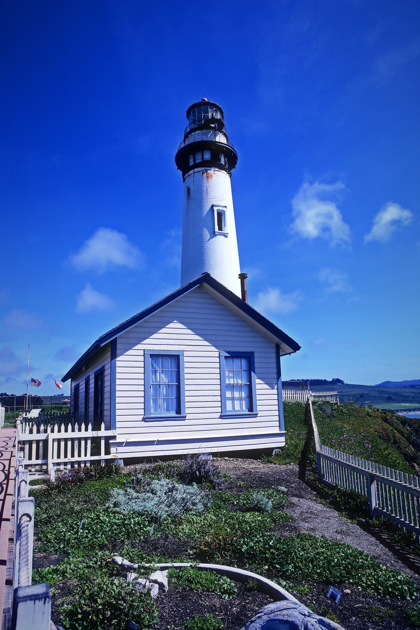 White lighthouse rising behind a white clapboard keeper's house under a deep blue sky.