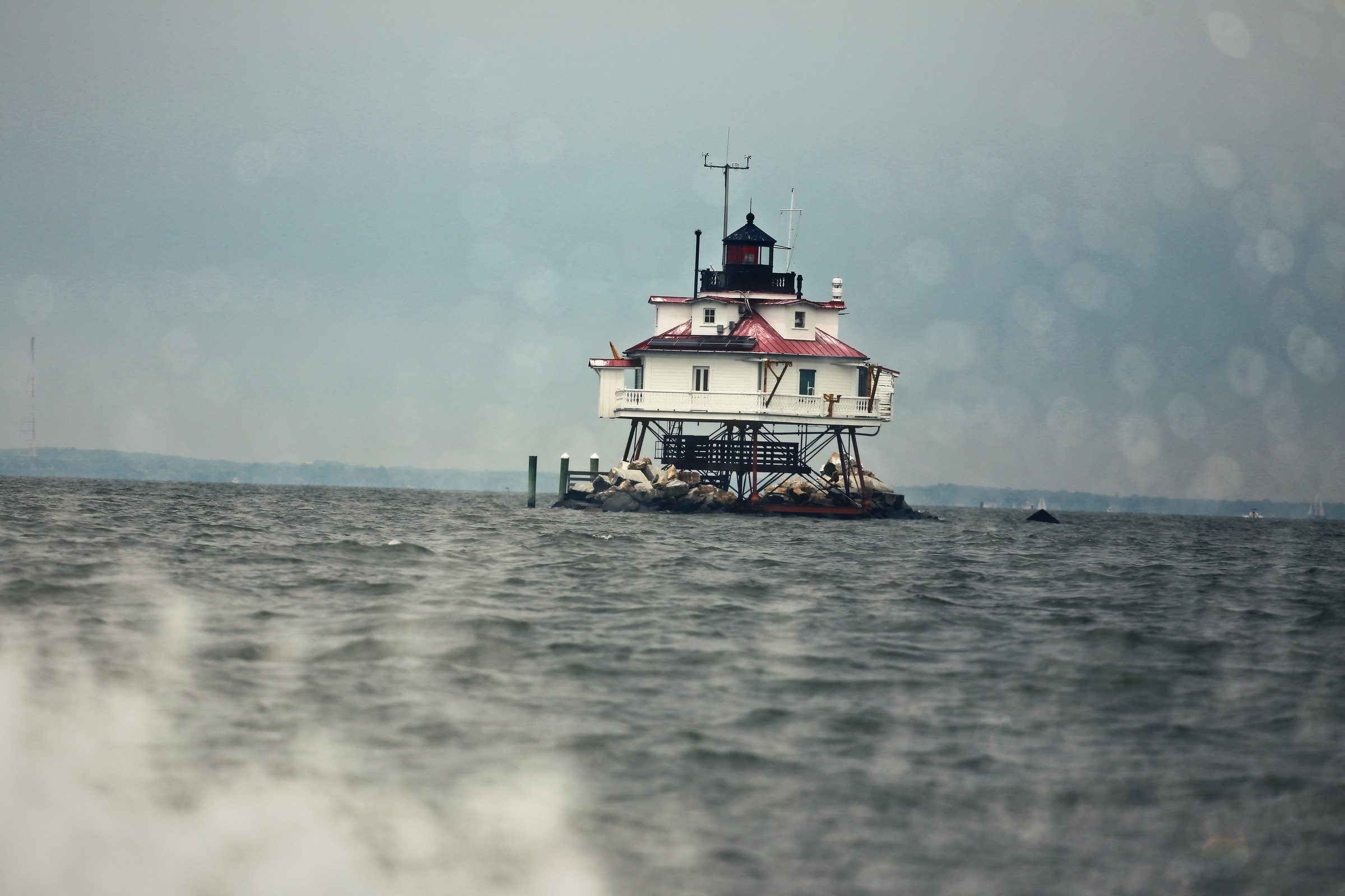 Hexagonal cottage lighthouse standing in open water behind wind-blown spray.