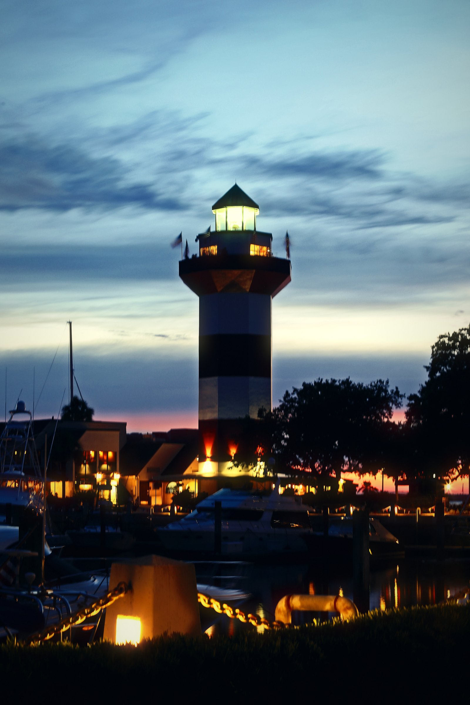 Striped lighthouse with its lamp on above a marina as the last sunset color fades.