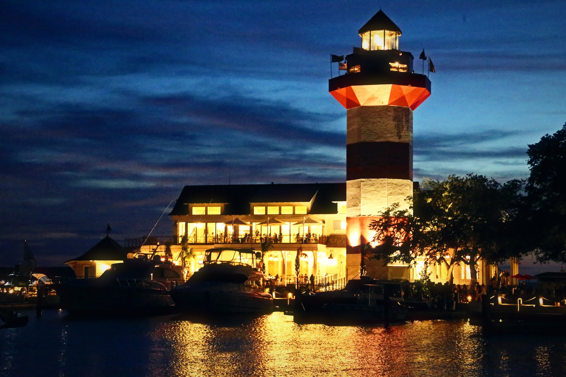 Lit lighthouse and harborfront buildings reflected in calm water at night.