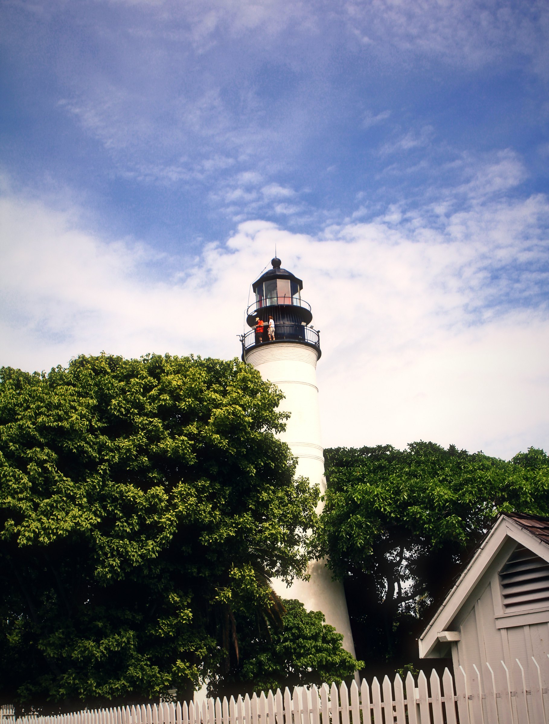 White lighthouse rising over green trees beyond a white picket fence and cottage roof.