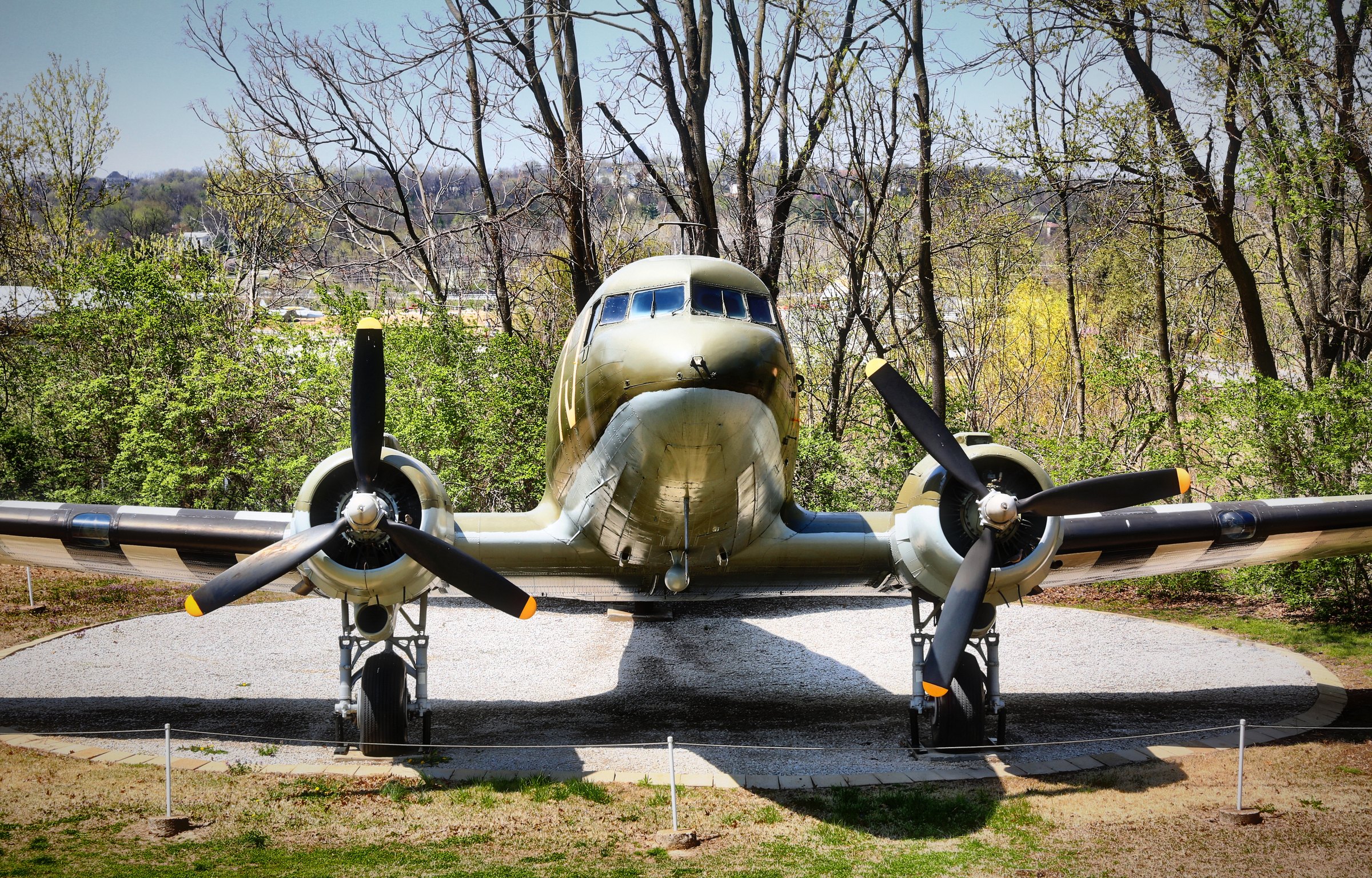 A vintage twin-propeller airplane on display, nose-on, framed by bare spring trees.