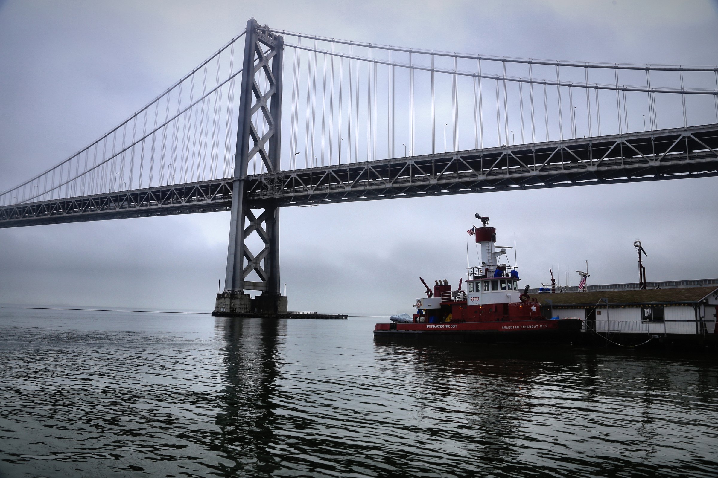 A red fireboat moored beneath a long suspension bridge on still grey water.