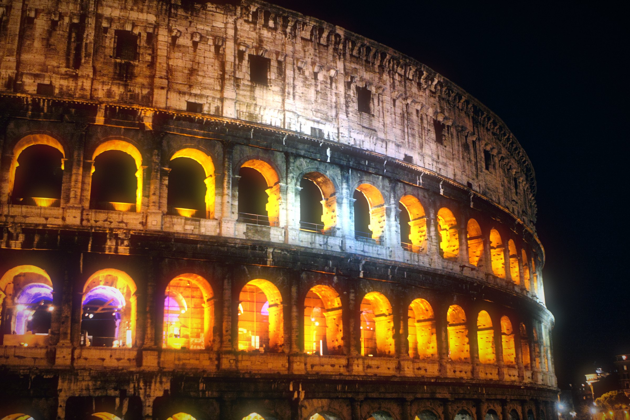The Colosseum at night, its arches glowing amber and violet.