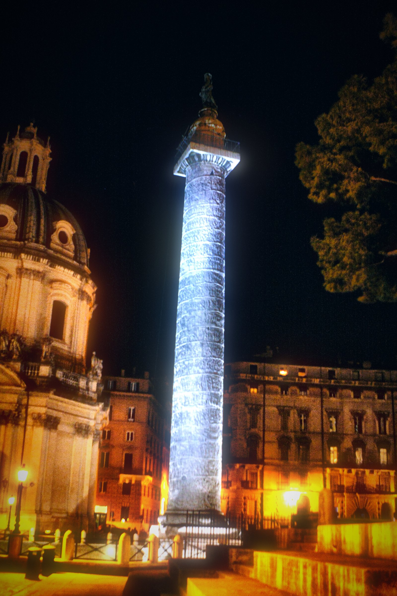 Trajan's Column lit white at night beside a church dome.