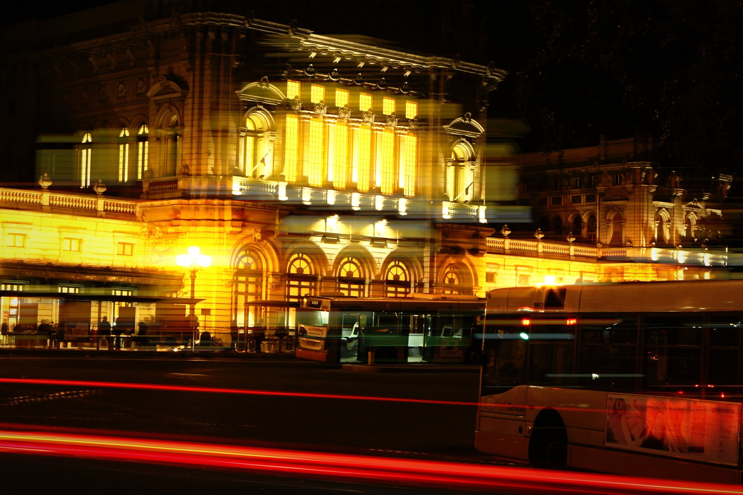 Floodlit building at night with long red light trails and a passing bus.