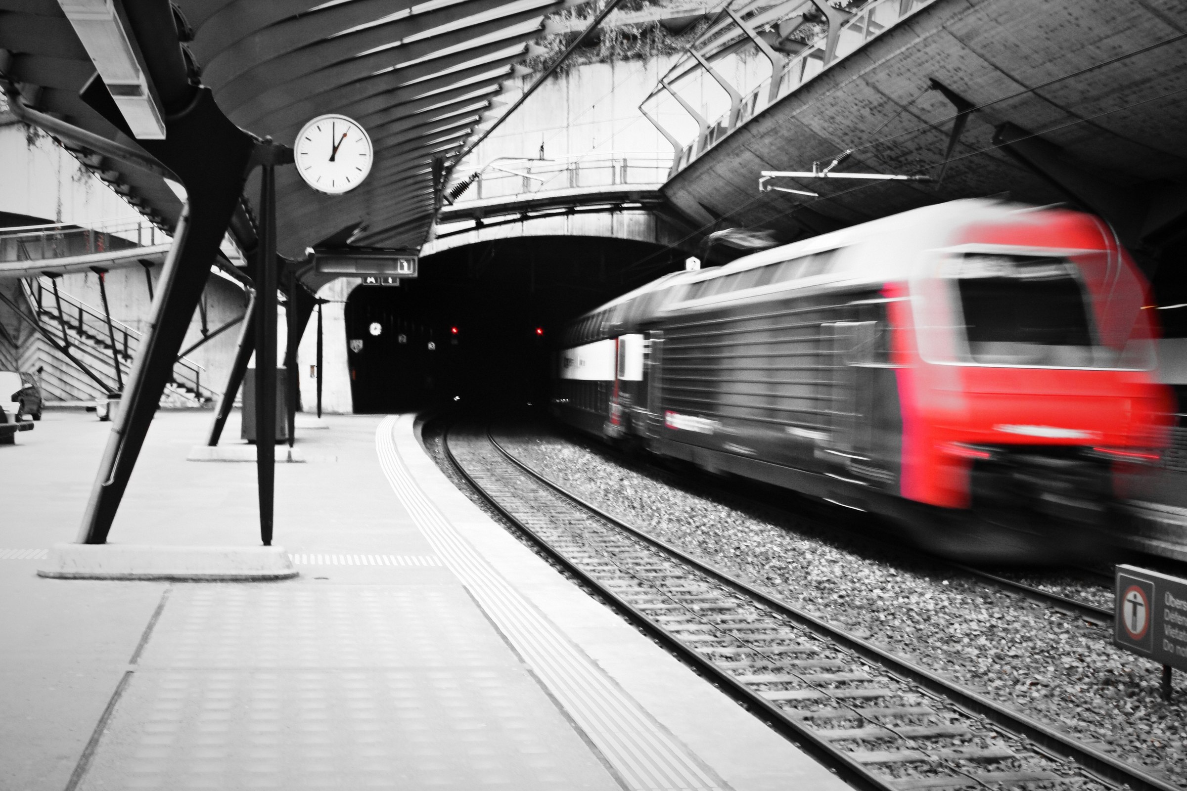 Black and white platform under a steel canopy with a speeding red locomotive in colour.