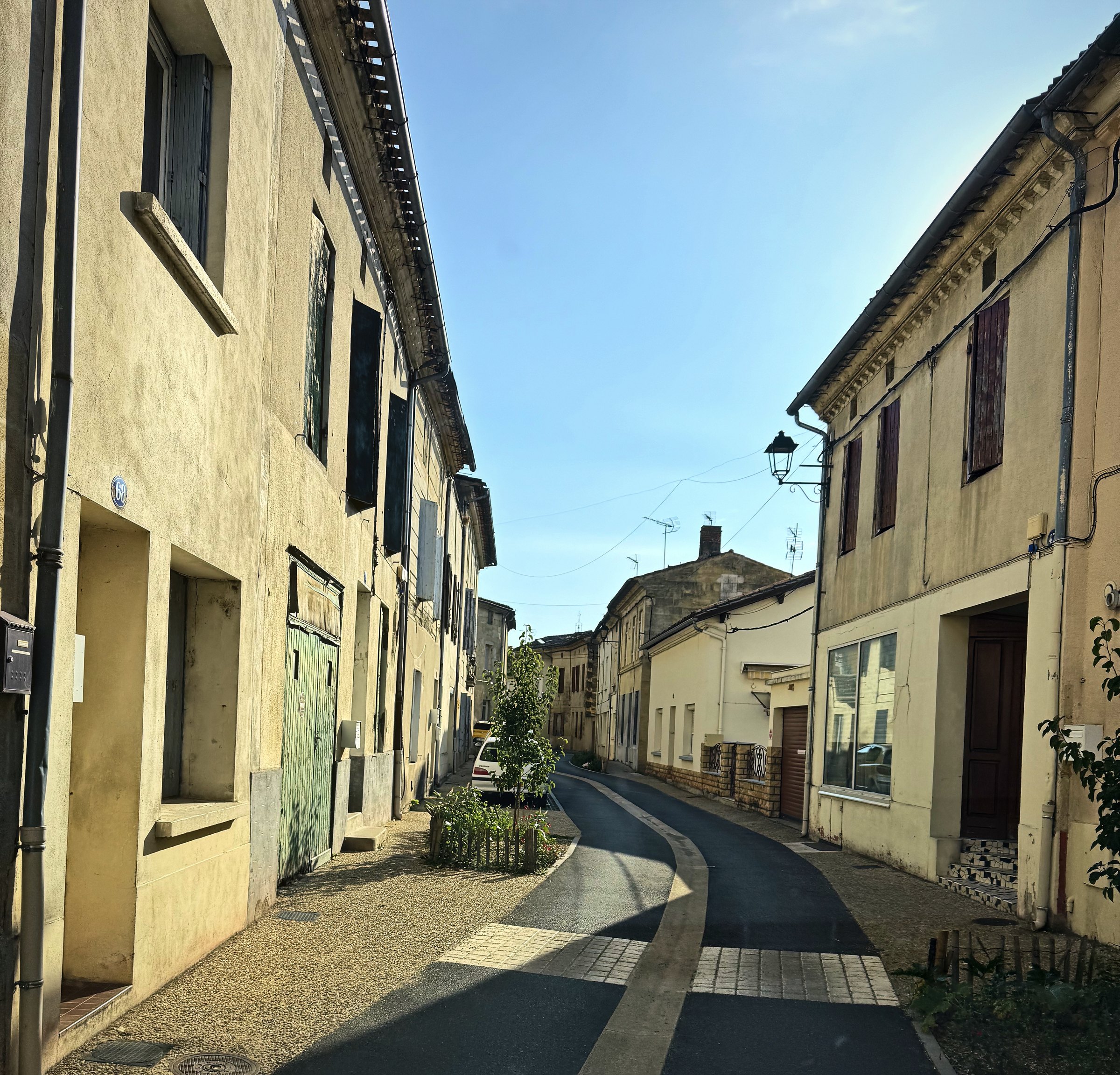 A quiet street of pale stone houses with shutters and a narrow pavement in early sun.