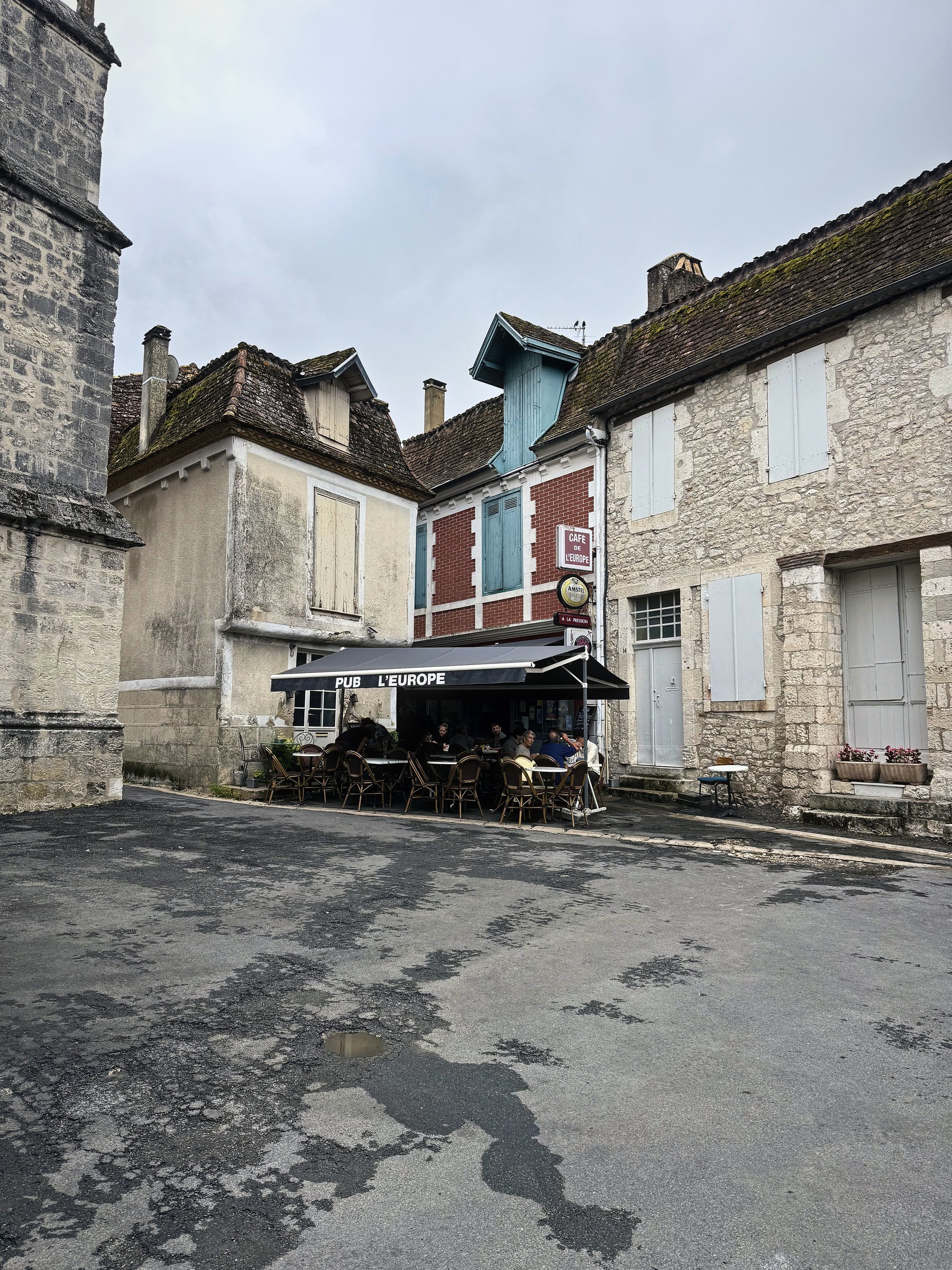 A bend of an old stone village street with shuttered houses and a café awning.