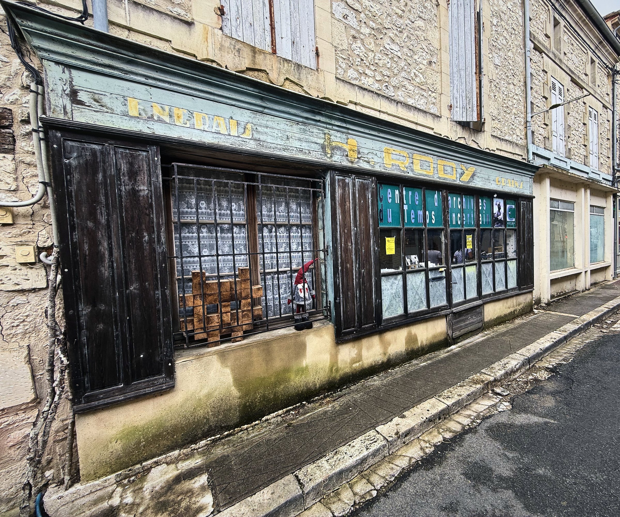 A long, faded wooden shopfront running along an old stone building on a village street.