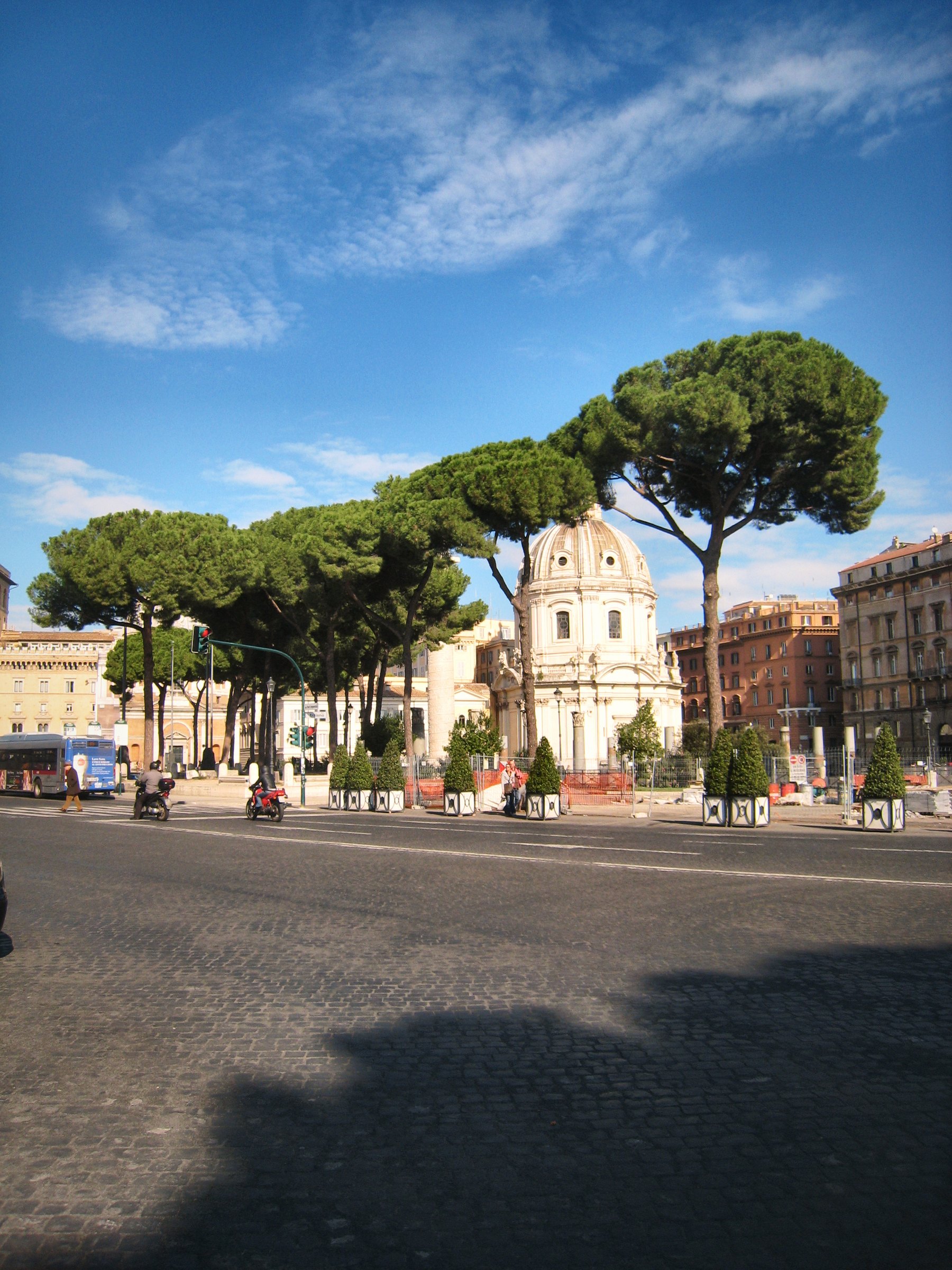 Umbrella pines over a Roman street and a church facade under blue sky.