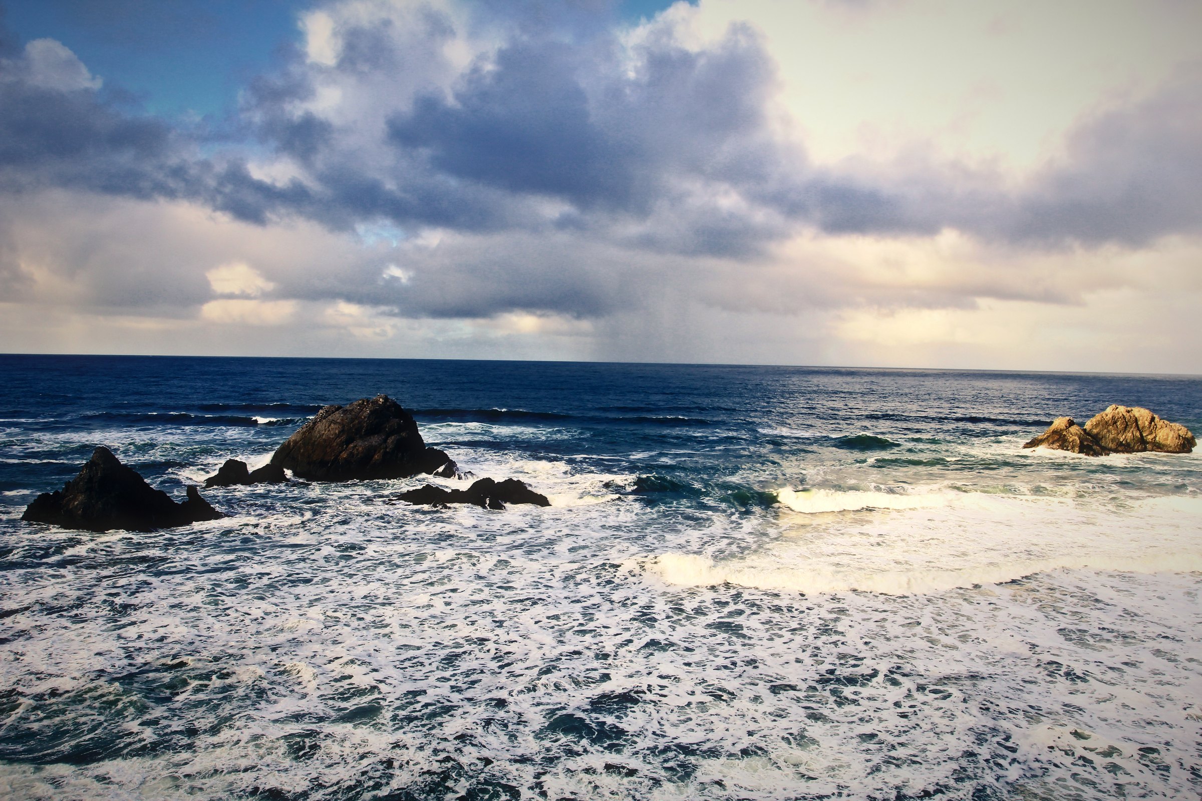 Low sea stacks surrounded by white foam under heavy clouds and a bright horizon.