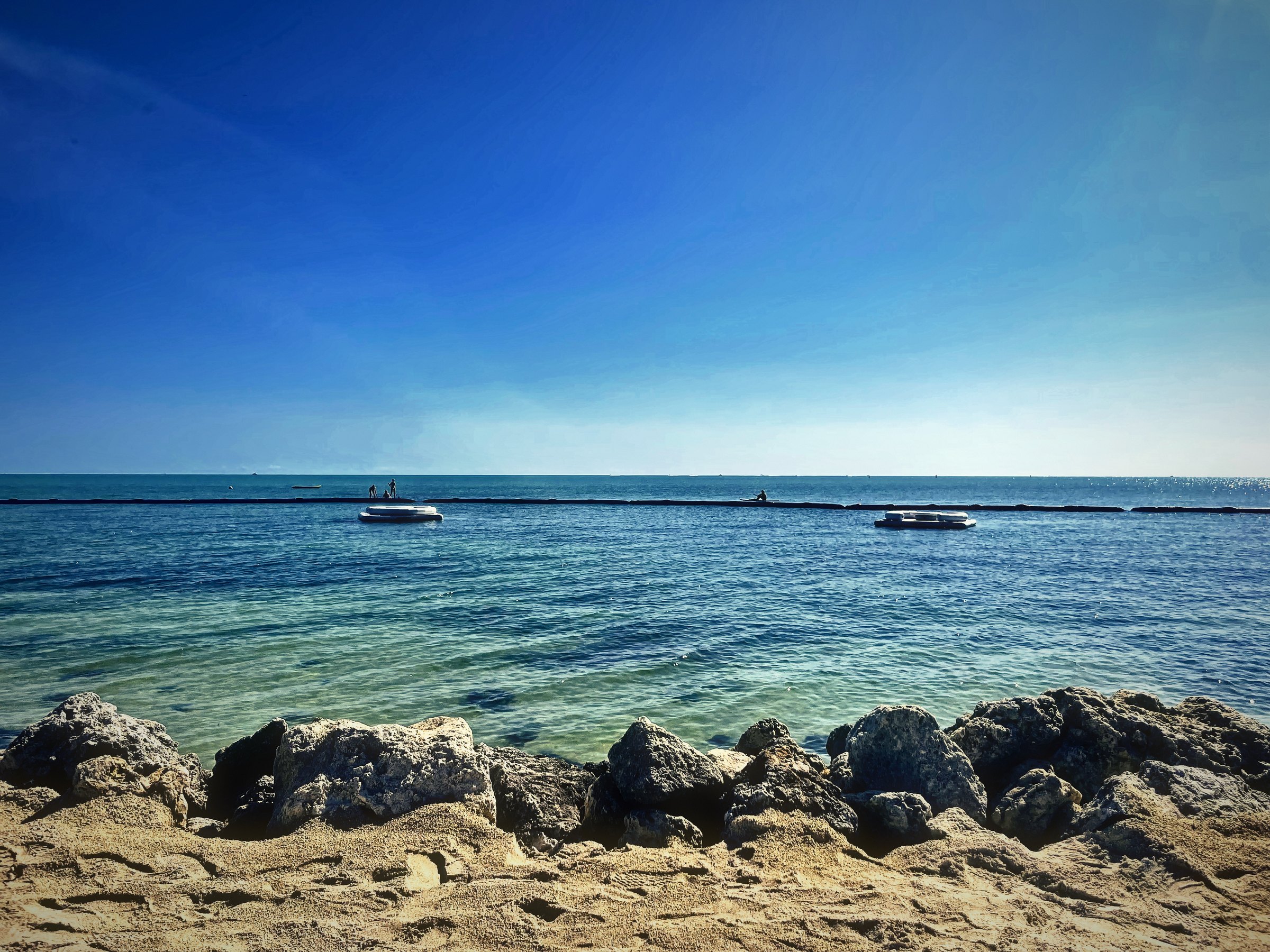 A line of rough boulders in front of calm turquoise water and a pale horizon.
