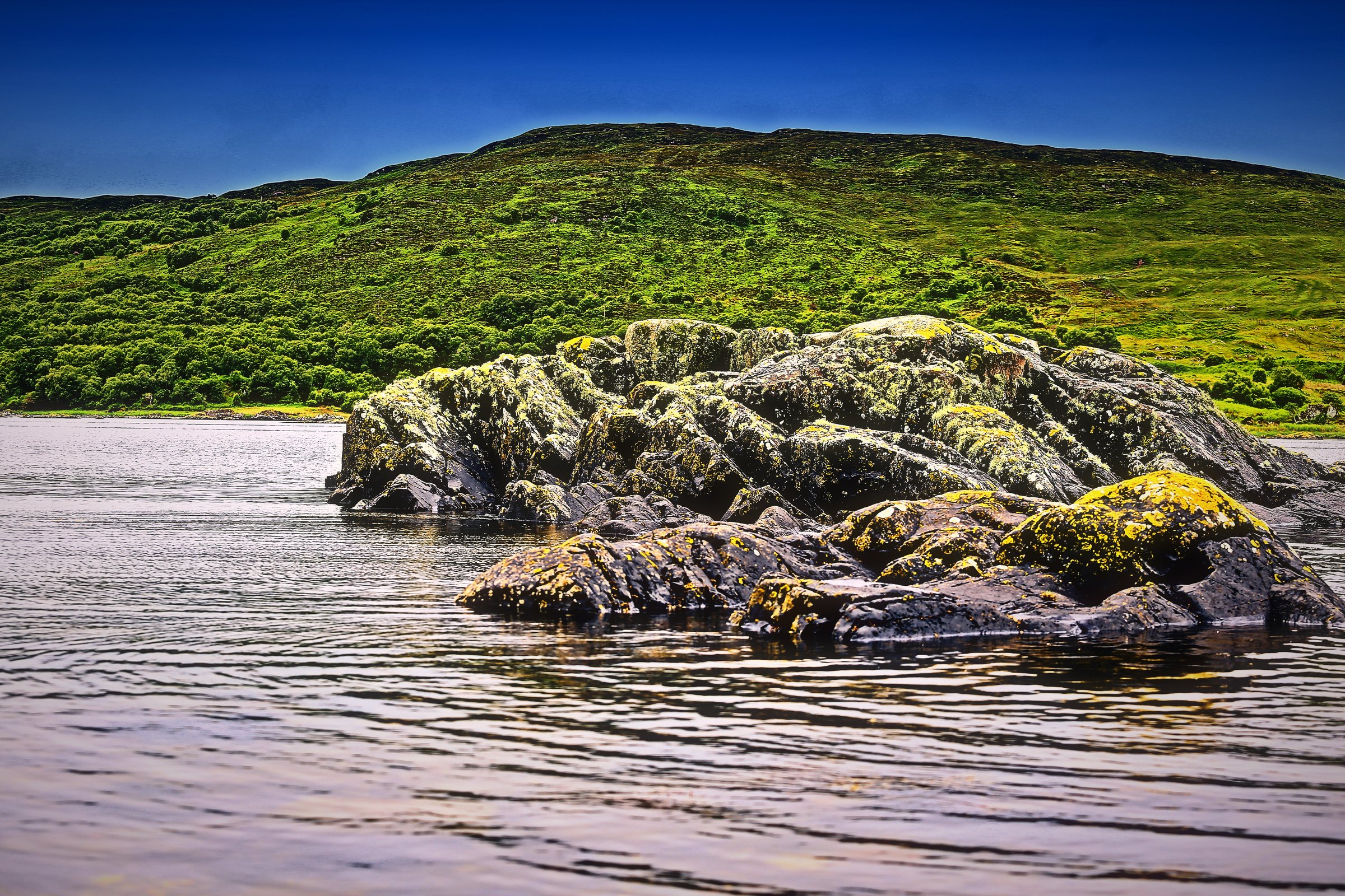 Lichen-covered rocks on a shallow silver shore below a green hillside.