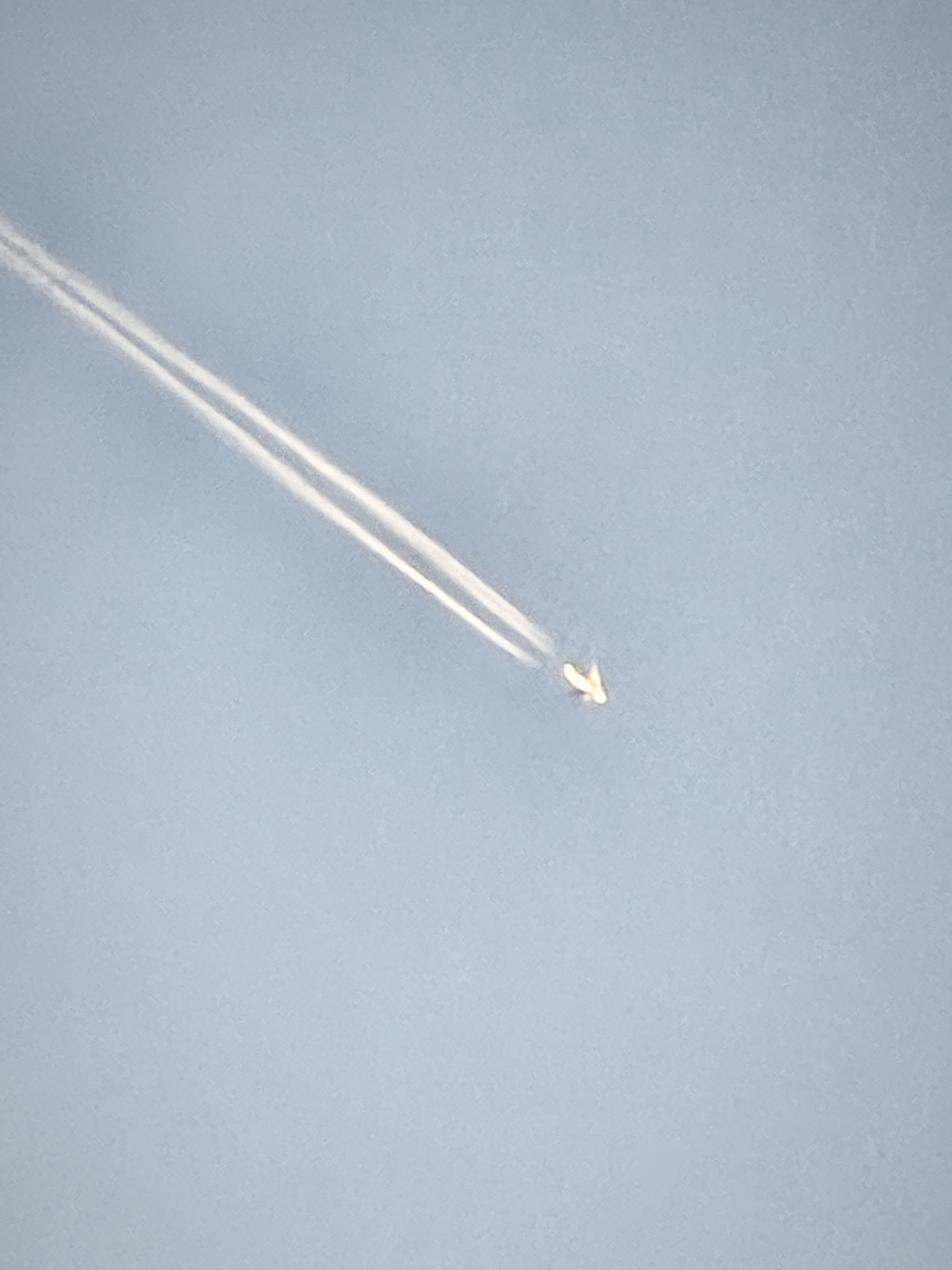 A high jet and its contrail cutting diagonally across a pale sky.